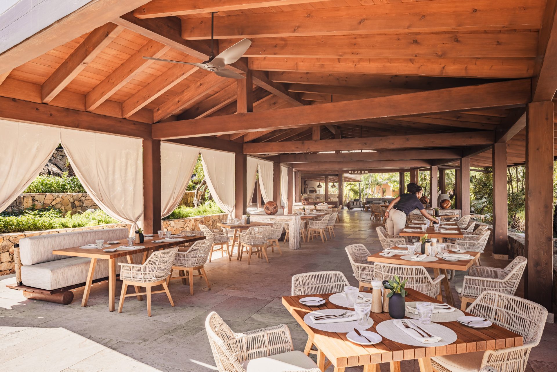 The Beach Club is an outdoor restaurant with wooden beams and ceiling, featuring white wicker chairs and wooden tables set for dining. Light streams through the open sides with white curtains, creating a bright, airy atmosphere. Lush greenery is visible outside.