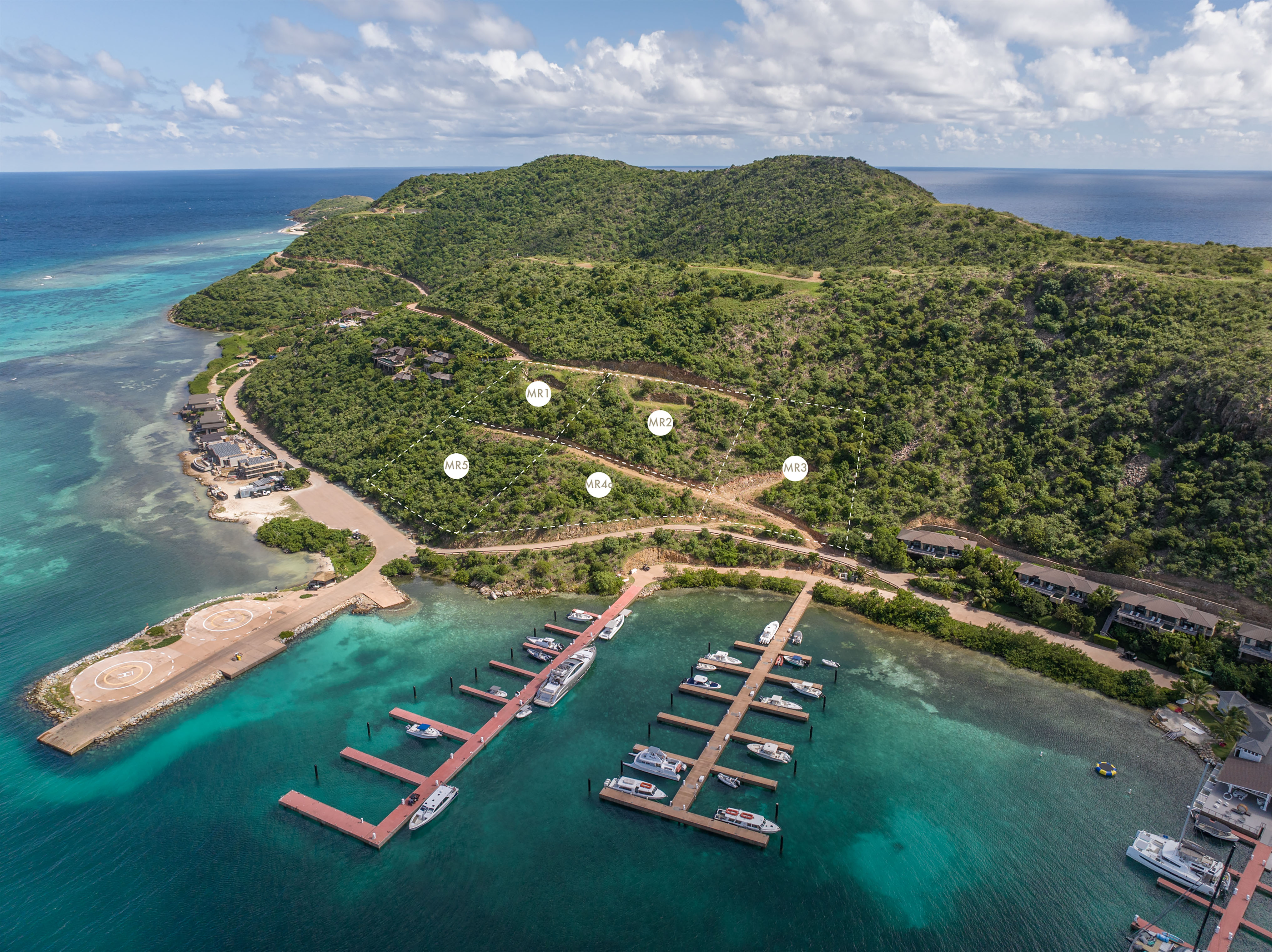 A scenic aerial view of a coastal area with a marina featuring several docks and boats. There are lush green hills and a prominent ridge in the background, a winding road, and various buildings scattered near the coastline. The turquoise waters of the sea surround the area.