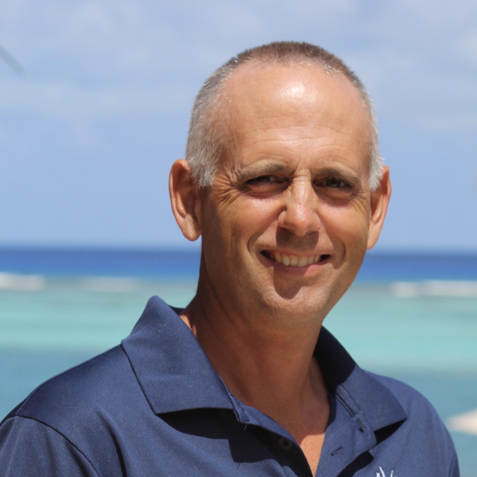 A smiling bald man with a short haircut, Mark Moore, stands outdoors in front of a backdrop of clear blue skies and a turquoise ocean. He is wearing a navy blue polo shirt.