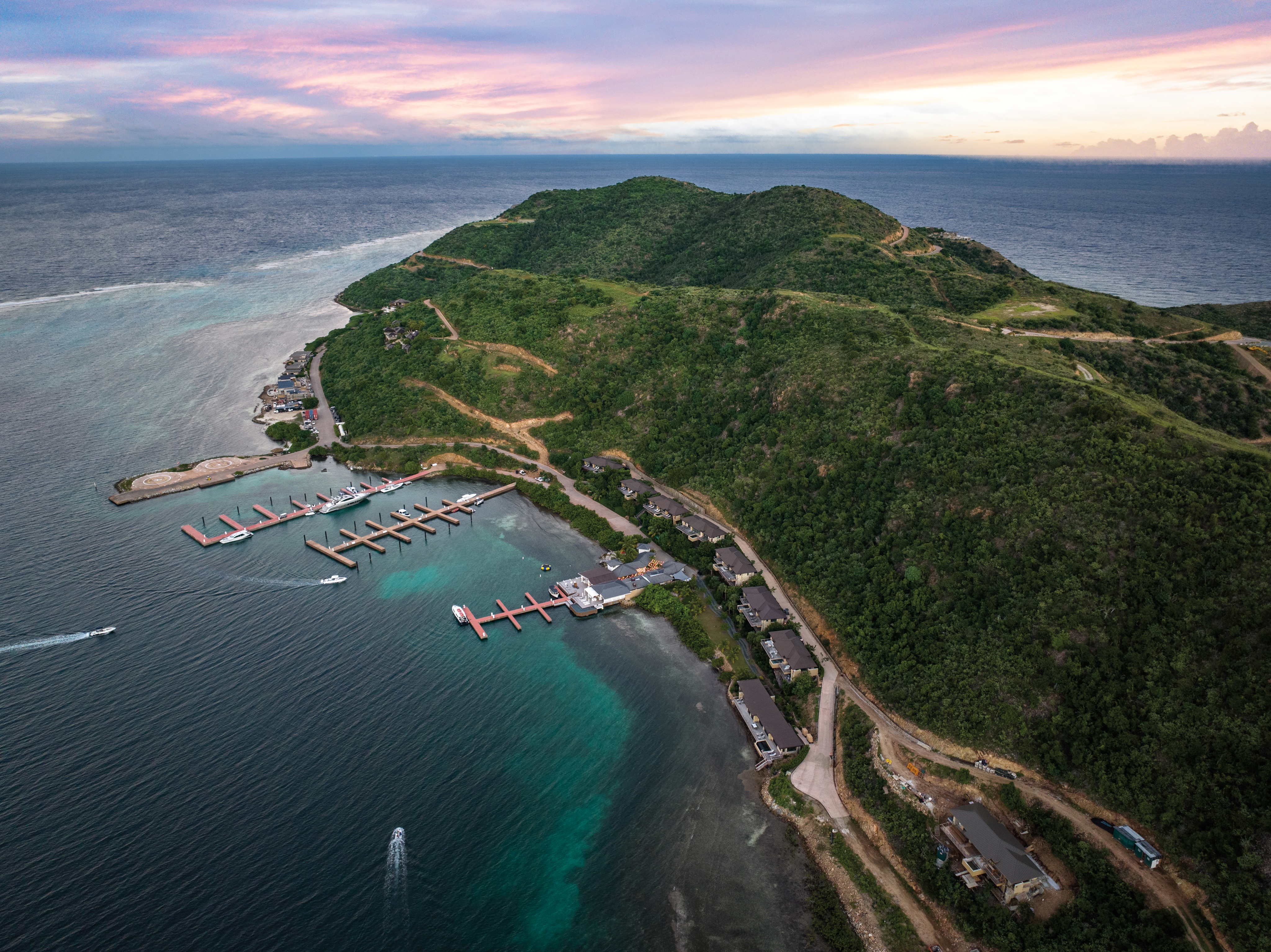 Aerial view of Marina Ridge's coastal area with lush green hills and vibrant blue waters. The coastline features several piers extending into the water and scattered buildings along the shore. The sky displays pastel shades of pink and purple, suggesting sunset or sunrise.