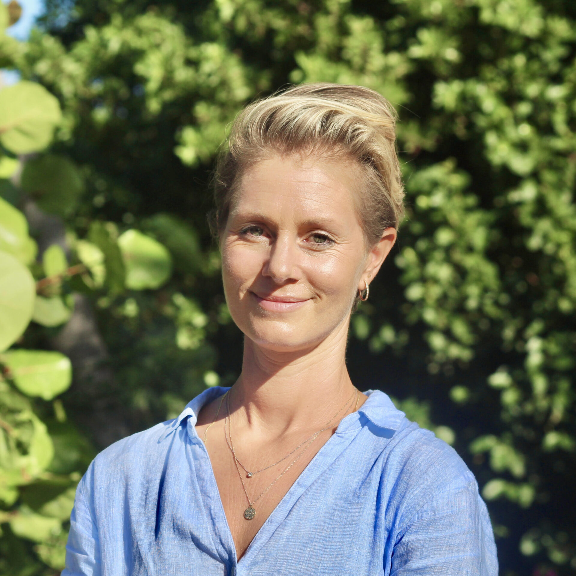 Emily Oakes, a woman with short blonde hair, stands outdoors in front of green foliage. She wears a light blue blouse and smiles gently at the camera. The bright lighting suggests a sunny day. Emily adorns two delicate necklaces and small hoop earrings.