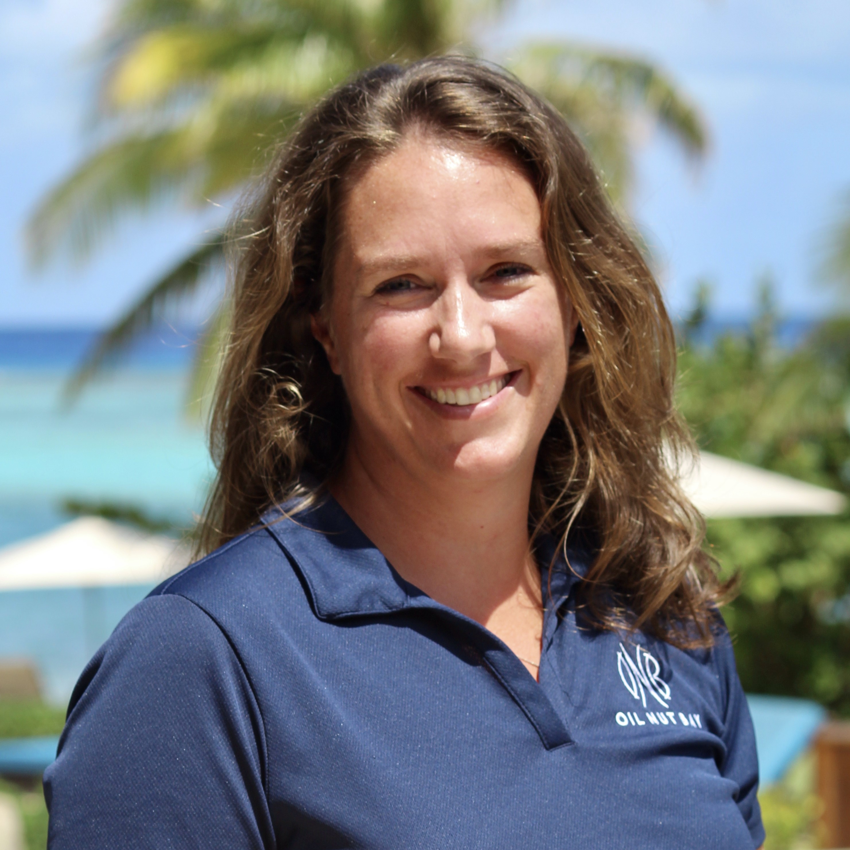 Sarah Moore, a woman with wavy brown hair, smiles at the camera. She is wearing a blue polo shirt with a logo on it, standing outdoors with palm trees and a bright blue ocean in the background on a sunny day.