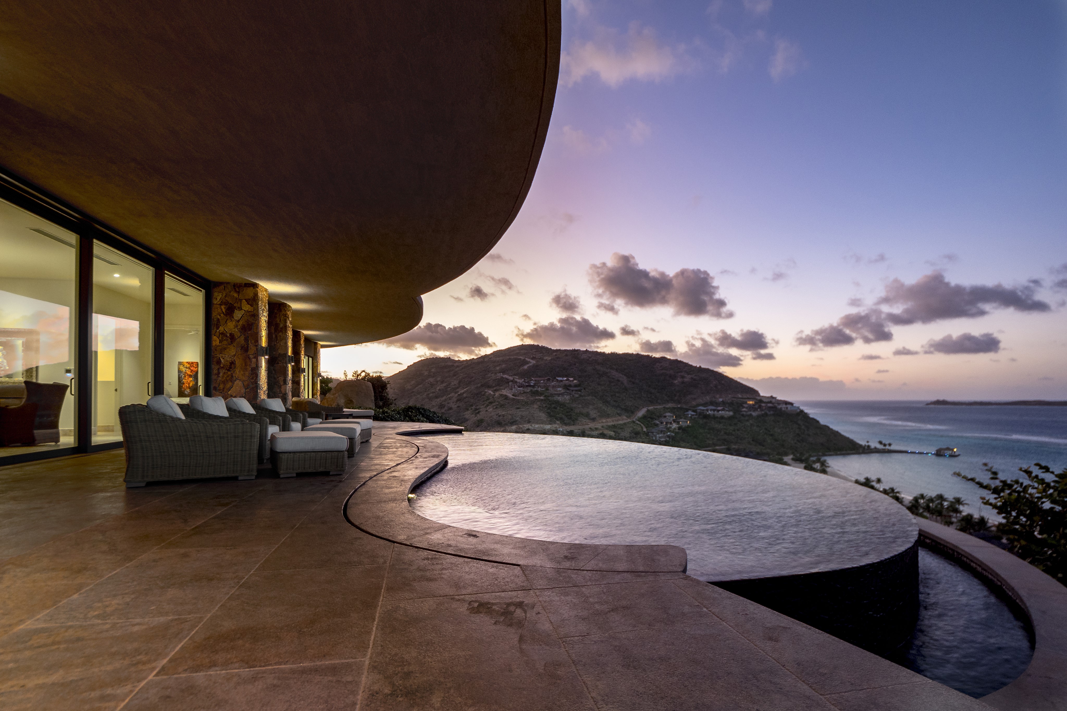 A luxury patio features three cushioned lounge chairs facing an infinity pool that seamlessly blends into the stunning view of the ocean and surrounding hilly landscape at sunset. The sky is filled with soft clouds and colors from the setting sun—ideal for enjoying while discussing management services for your BVI property.