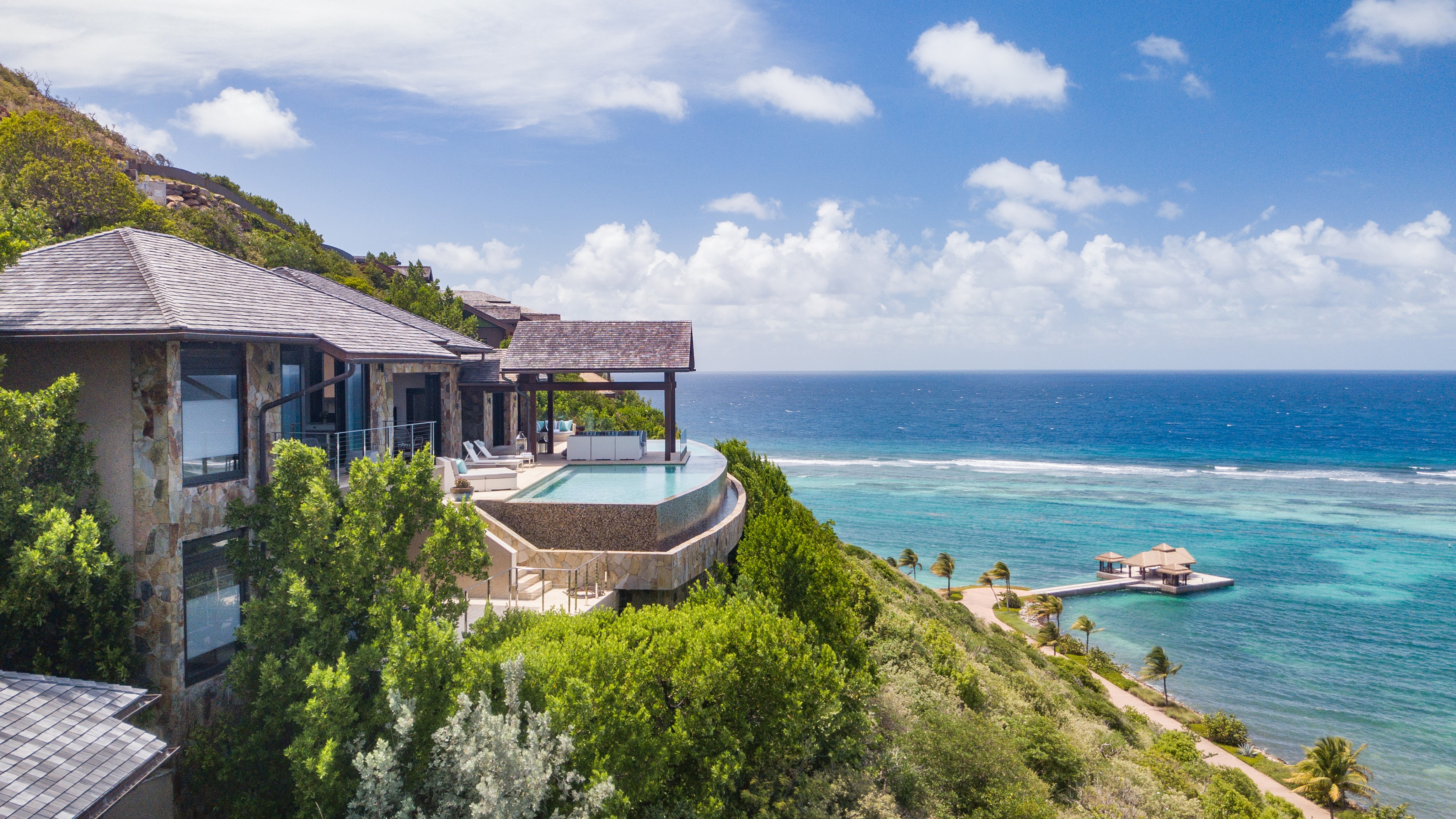 A luxurious seaside villa with an infinity pool overlooks the azure ocean and lush green hillside. The expansive view includes a distant dock with a hut on the right. The sky is clear with scattered clouds, and palm trees dot the coastline below—perfect for enjoying seamless management services for your BVI property.