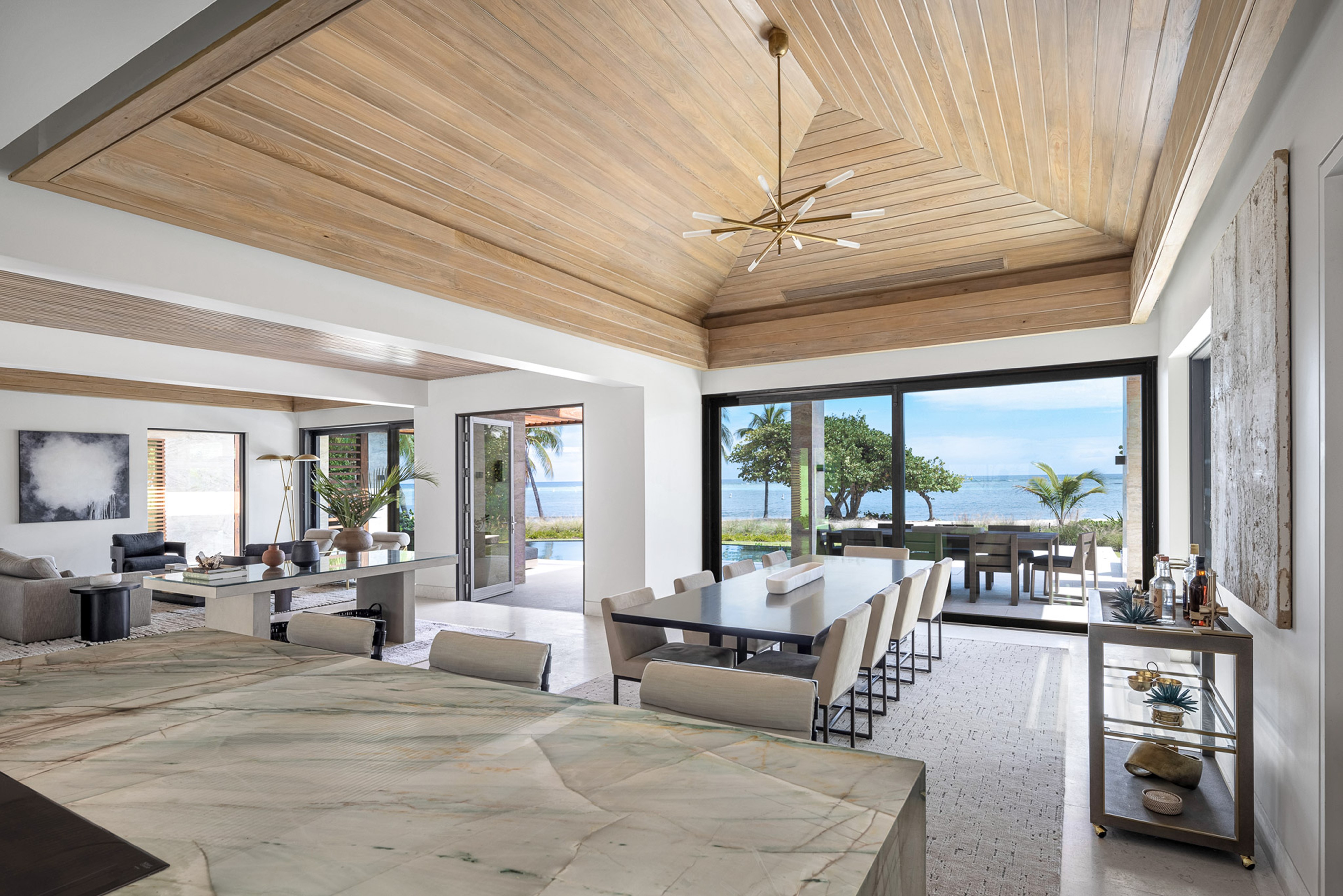 A modern open-concept living space with a wooden ceiling and large windows offering an ocean view. The room features a dining area with a long table and cream-colored chairs, a sitting area with sofas, and a kitchen island in the foreground. The space is decorated with plants, artwork, and provides seamless management services for your BVI property.