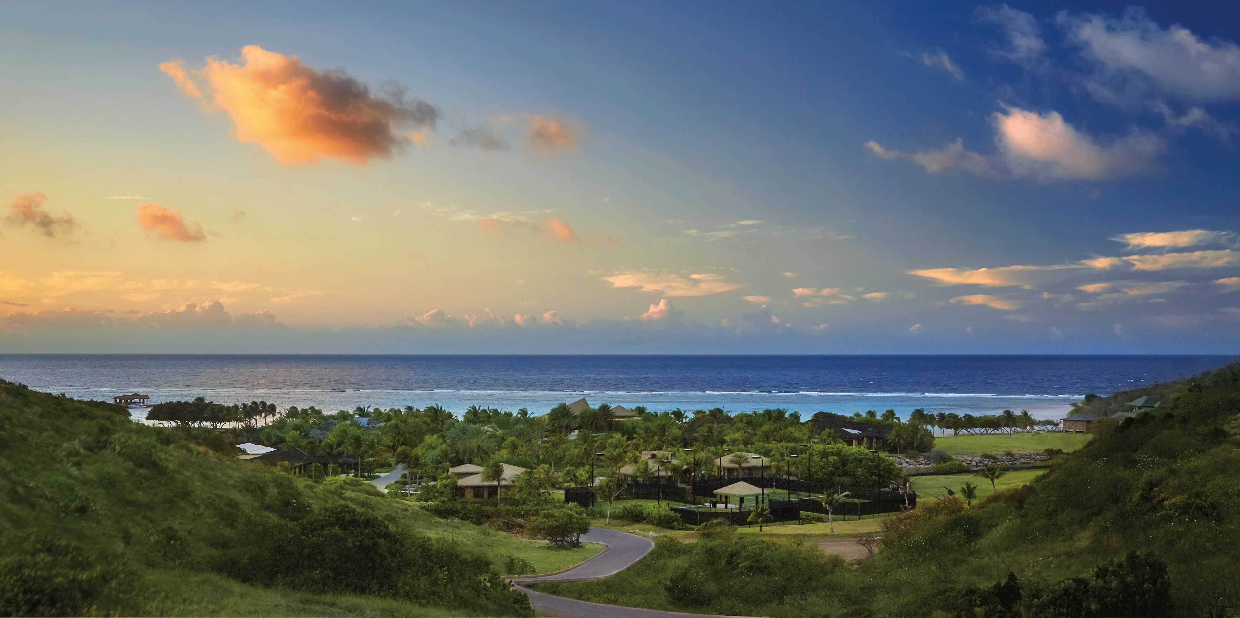 A scenic coastal landscape featuring lush green hillsides, a winding road, scattered buildings, and a vibrant blue ocean under a partly cloudy sky at sunset. The sky transitions from a warm orange near the horizon to a deep blue higher up—perfect for those seeking management services for your BVI property.