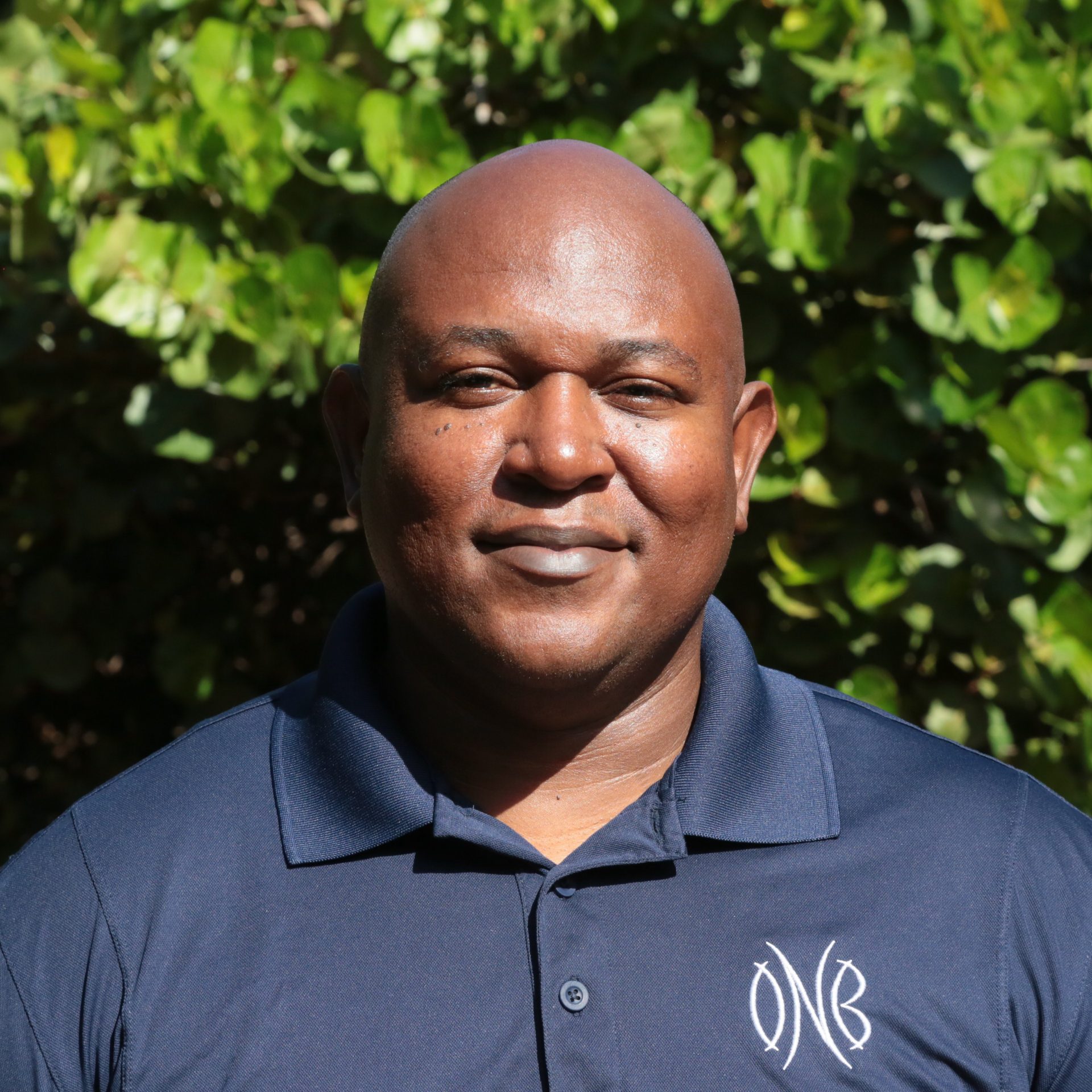 A person smiling and standing outdoors with greenery in the background. Vinod is wearing a dark blue polo shirt with a monogram on the left side of the chest. The individual has a shaved head and is looking directly at the camera.