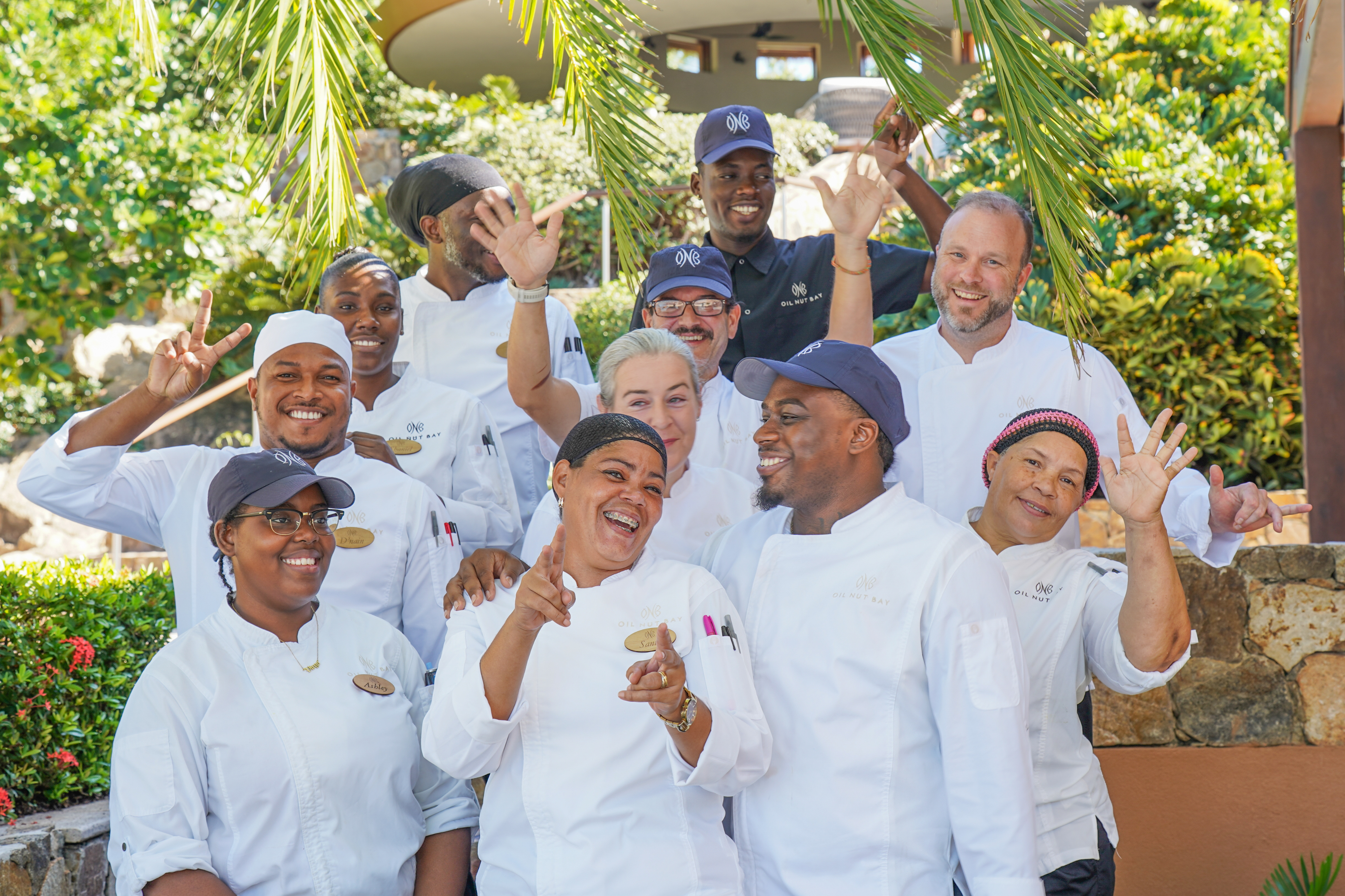 A group of chefs in white uniforms and hats poses together outdoors, smiling and making cheerful gestures. Some chefs are showing peace signs, while others are giving thumbs up. Greenery and part of a building are visible in the background, showcasing vibrant careers in the BVI's culinary scene.