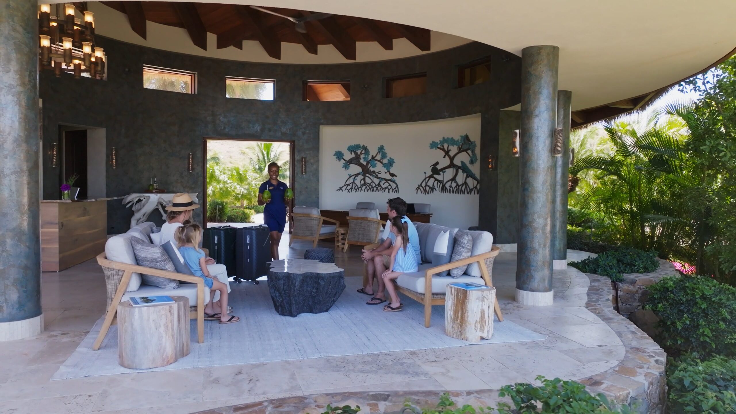 An outdoor reception area under a circular pavilion with a wooden ceiling, stone pillars, and a marble floor sets the stage for an exquisite arrival experience. A staff member assists a family seated on wooden chairs around a central stone table. The background features mural art and garden views.