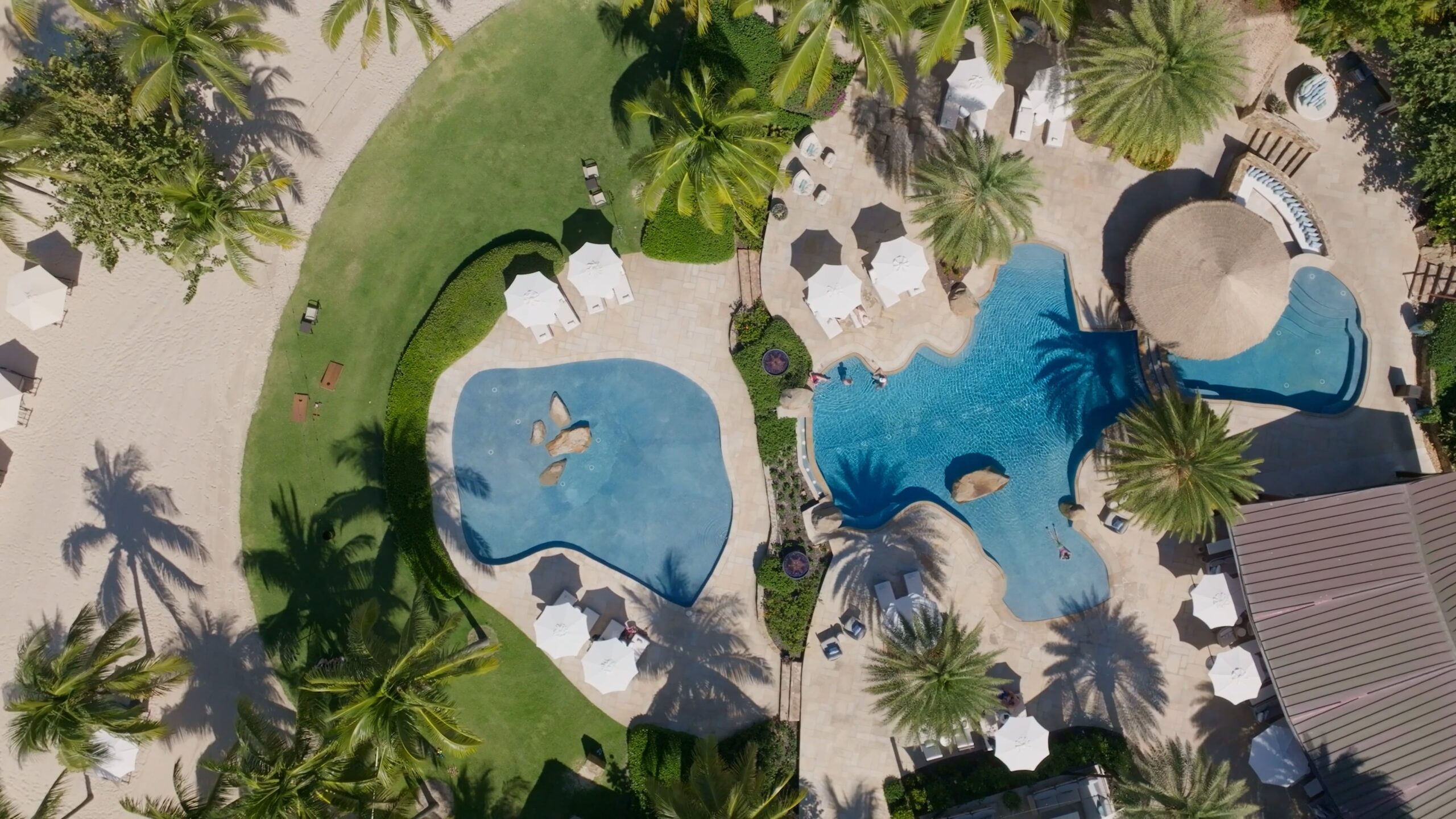 Aerial view of two adjacent pools surrounded by palm trees and lounge chairs with umbrellas at a beach club. One pool has a curvy shape and dual islands, while the other features a smaller island and an adjacent sandy area.