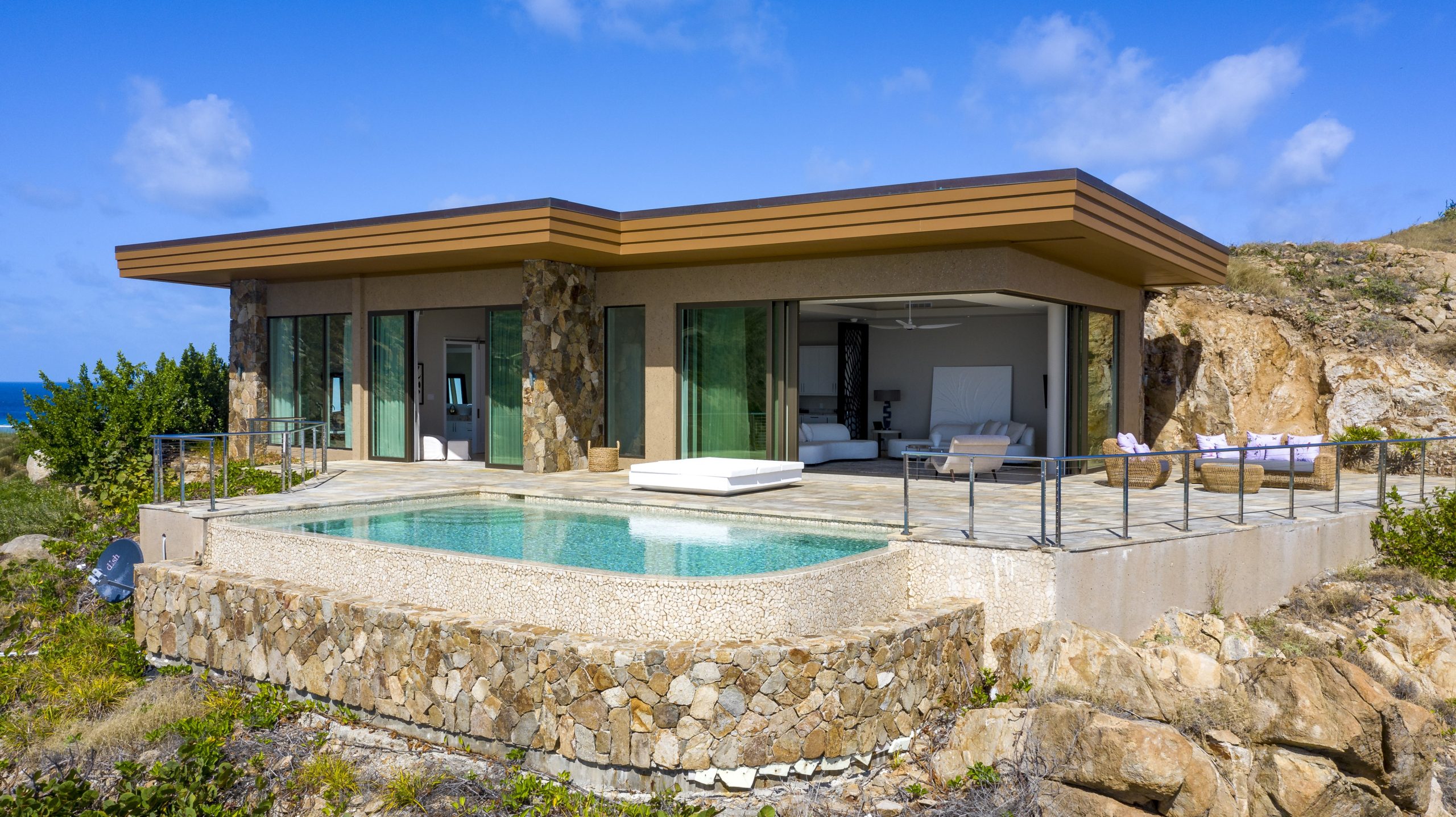 A modern, single-story luxury house with a flat roof is built into a rocky hillside. Boasting large glass windows and a spacious patio with lounge chairs, it resembles a cliffside penthouse complete with an infinity pool overlooking the ocean under a clear blue sky.