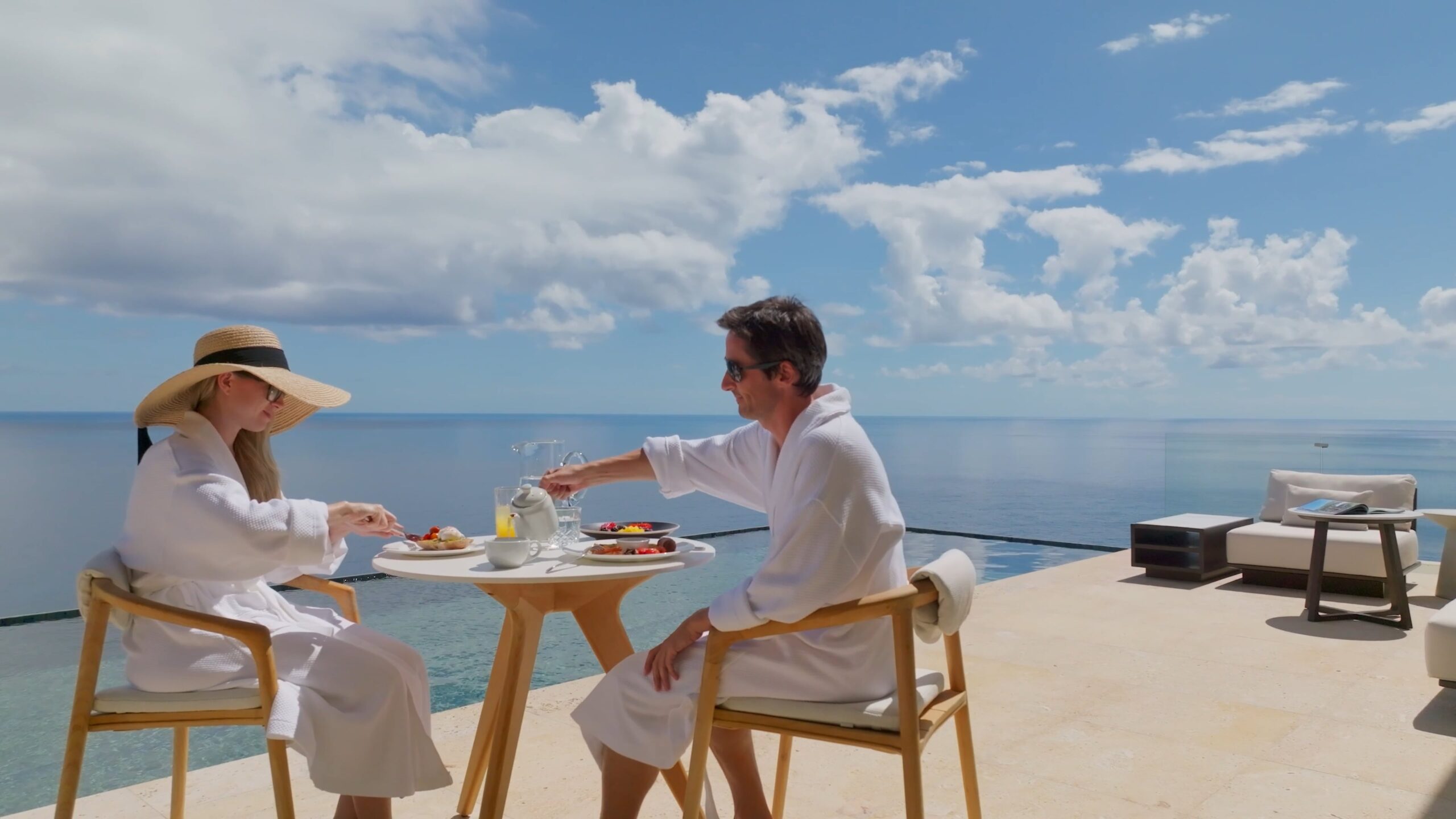 A couple wearing white bathrobes and sunglasses sit at a small table on a luxurious terrace overlooking the ocean. They are dining under a clear, blue sky with scattered clouds, savoring a meal in this serene and relaxing setting. An infinity pool glistens in the background.