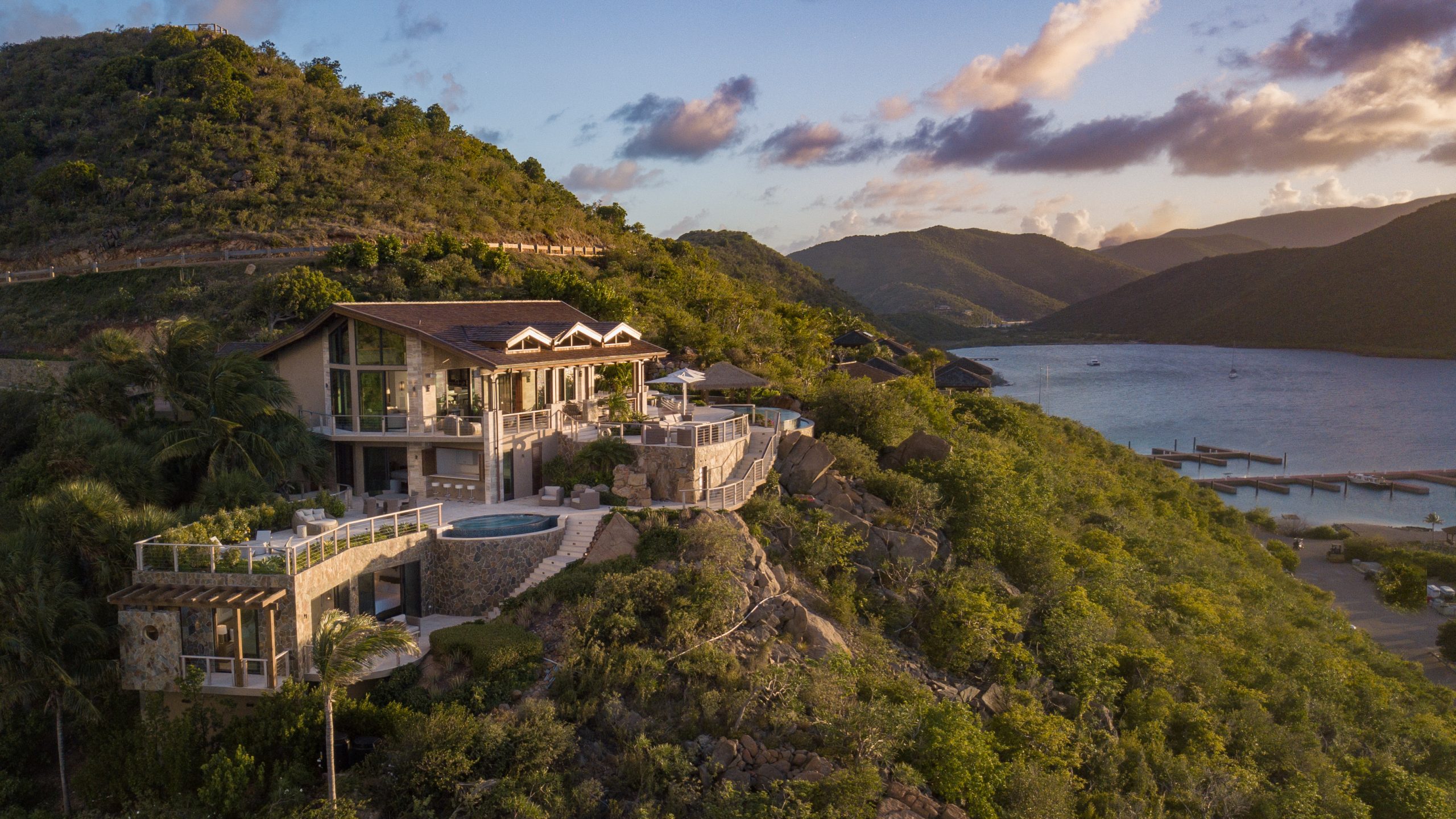 A luxurious hillside estate overlooking a serene bay with mountainous terrain in the background. The villa features multiple levels, expansive balconies, and a circular swimming pool, all bathed in the warm glow of the setting sun.