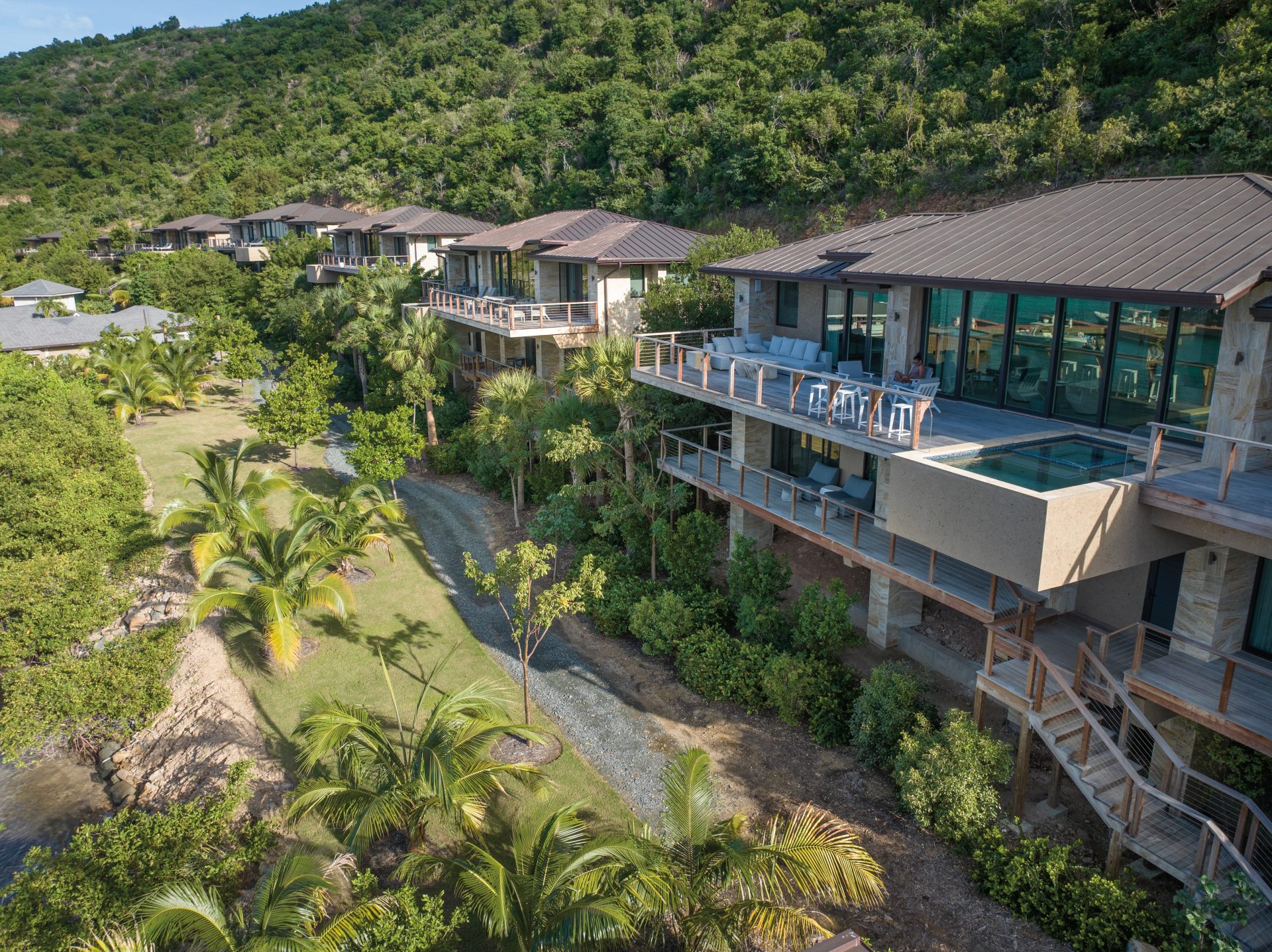 A series of luxurious hillside villas with large glass windows and balconies overlook a lush, green landscape with palm trees. The serene setting, akin to an exclusive neighborhood, includes a pathway winding through the vegetation and a view of distant mountains. The sky is clear, suggesting a sunny day.