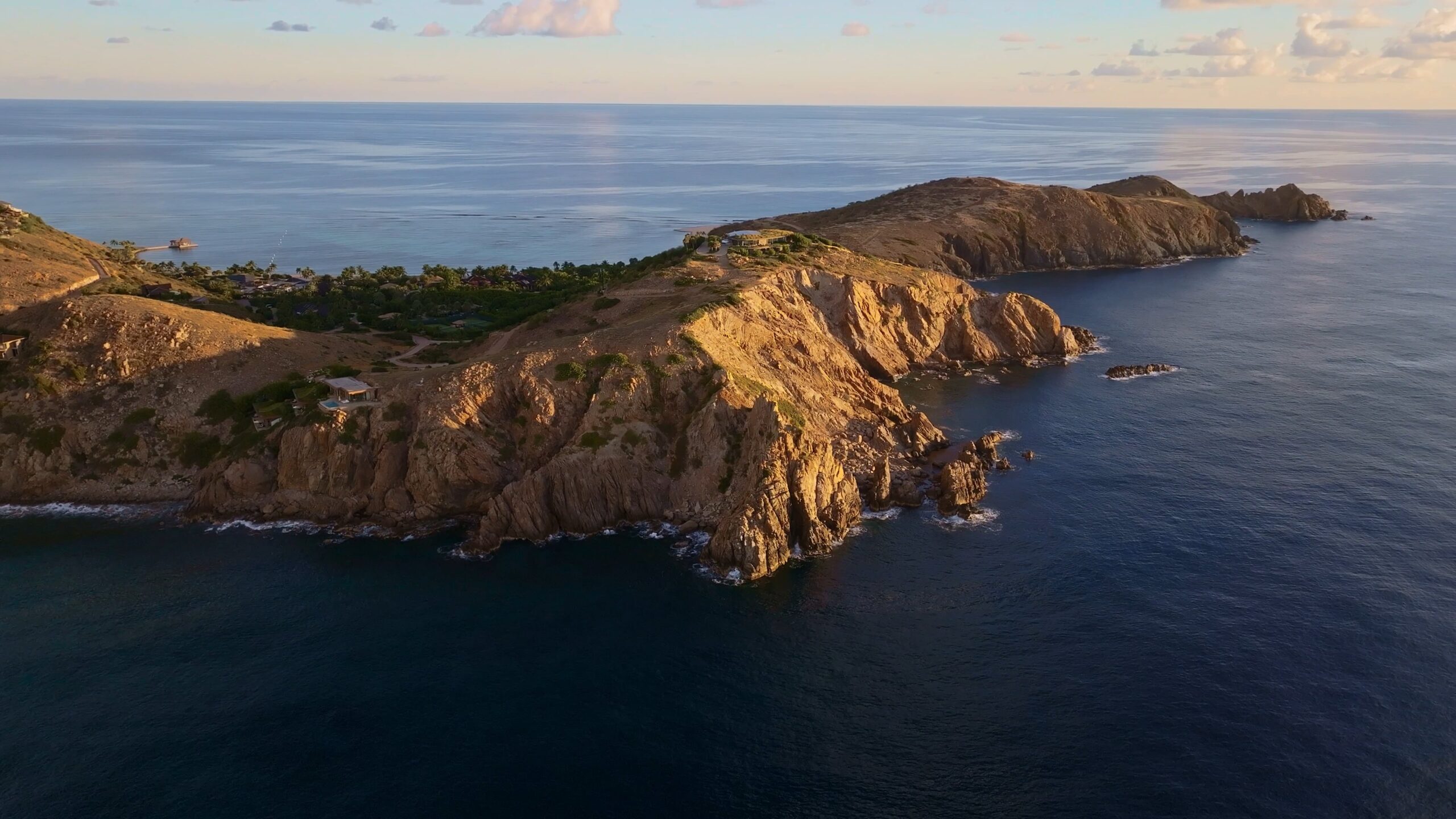 Aerial view of Oil Nut Bay's rocky coastal landscape jutting out into the ocean. The terrain features steep cliffs with patches of greenery on top. The sea is calm, reflecting the soft light of the setting sun. Scattered clouds dot the sky, enhancing the serene atmosphere near the resort.