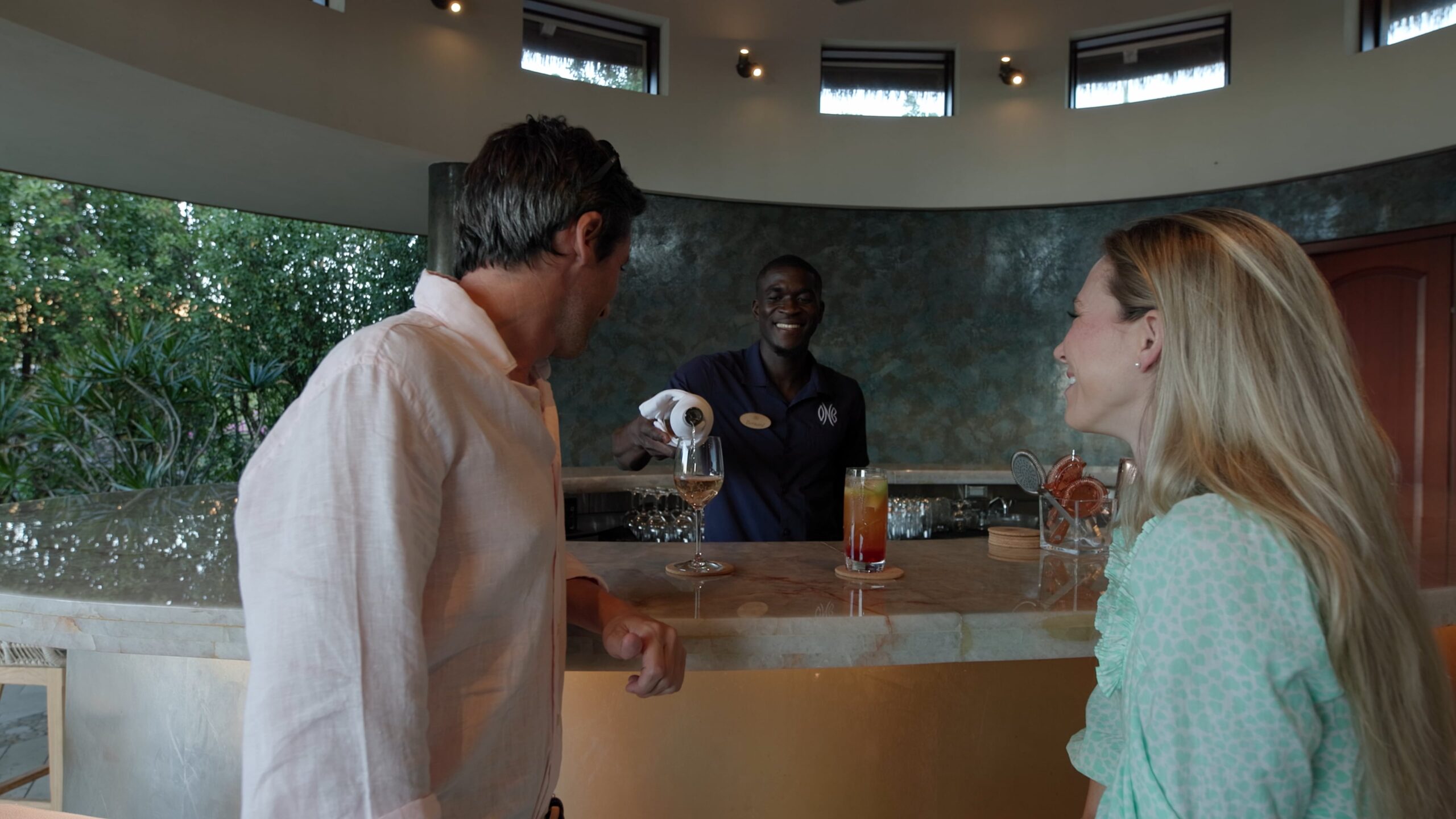 A man and woman stand at a stylish pavilion bar, smiling, as a bartender wearing a name tag and a dark shirt serves drinks. The bar counter displays mixed drinks and a wine glass. The background features a curved stone wall with ambient lighting and greenery visible through the windows.