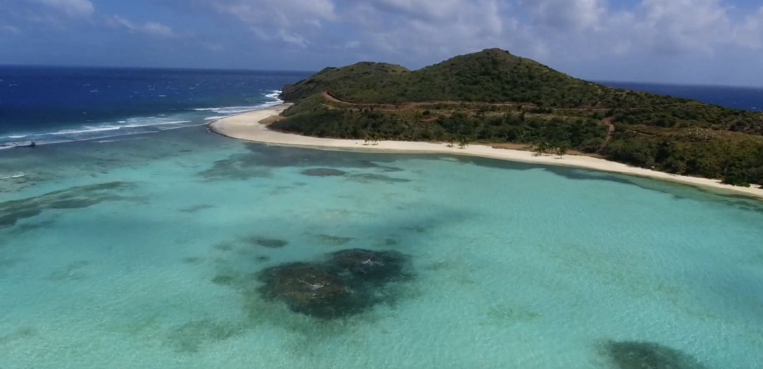 Aerial view of a tropical beach on a picturesque peninsula with clear turquoise water, sandy shores, and lush green hilly terrain in the background. The ocean waves gently crash along the shore under a partly cloudy sky, creating an idyllic neighborhood-like coastal scene.