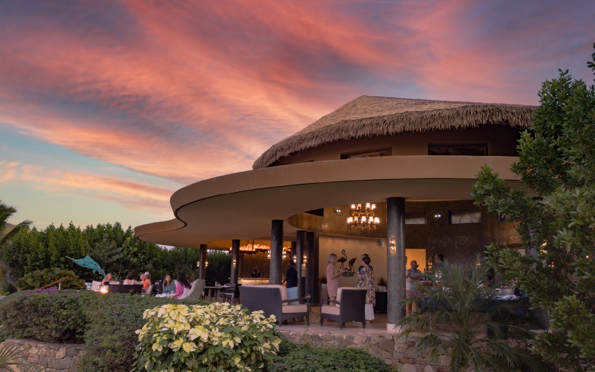 A luxurious, modern villa with a large curved patio is bustling with people enjoying a gathering by BVI Weddings & Events. The sky above displays a vibrant sunset with shades of orange and red. The lush greenery surrounds the property, adding to the serene ambiance.