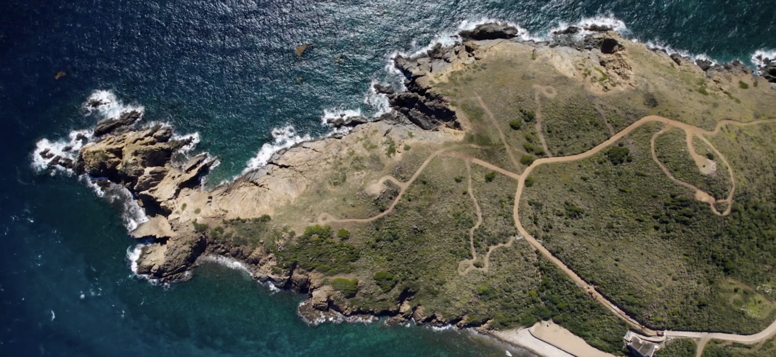 Aerial view of a rugged, rocky coastline with green vegetation and worn dirt paths winding across the terrain. Waves crash against the cliffs, and the deep blue and turquoise ocean surrounds this wildside neighborhood. A small sandy beach is visible at the bottom.