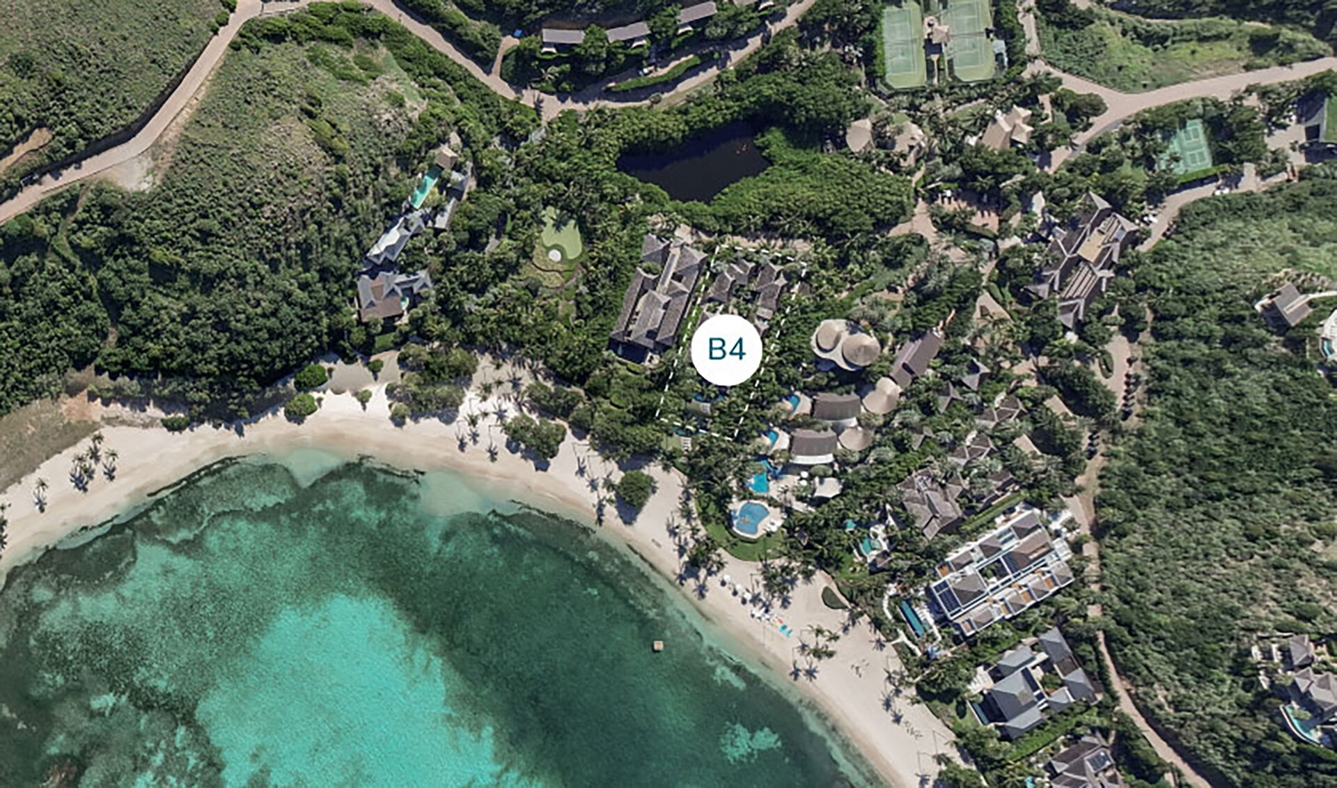 Aerial view of Nonamé Villa within a coastal resort area, featuring a mix of buildings and lush greenery. Several pools, pathways, and structures are clustered near the shoreline. The turquoise ocean is at the bottom of the image, with white sandy beaches leading up to the picturesque resort.