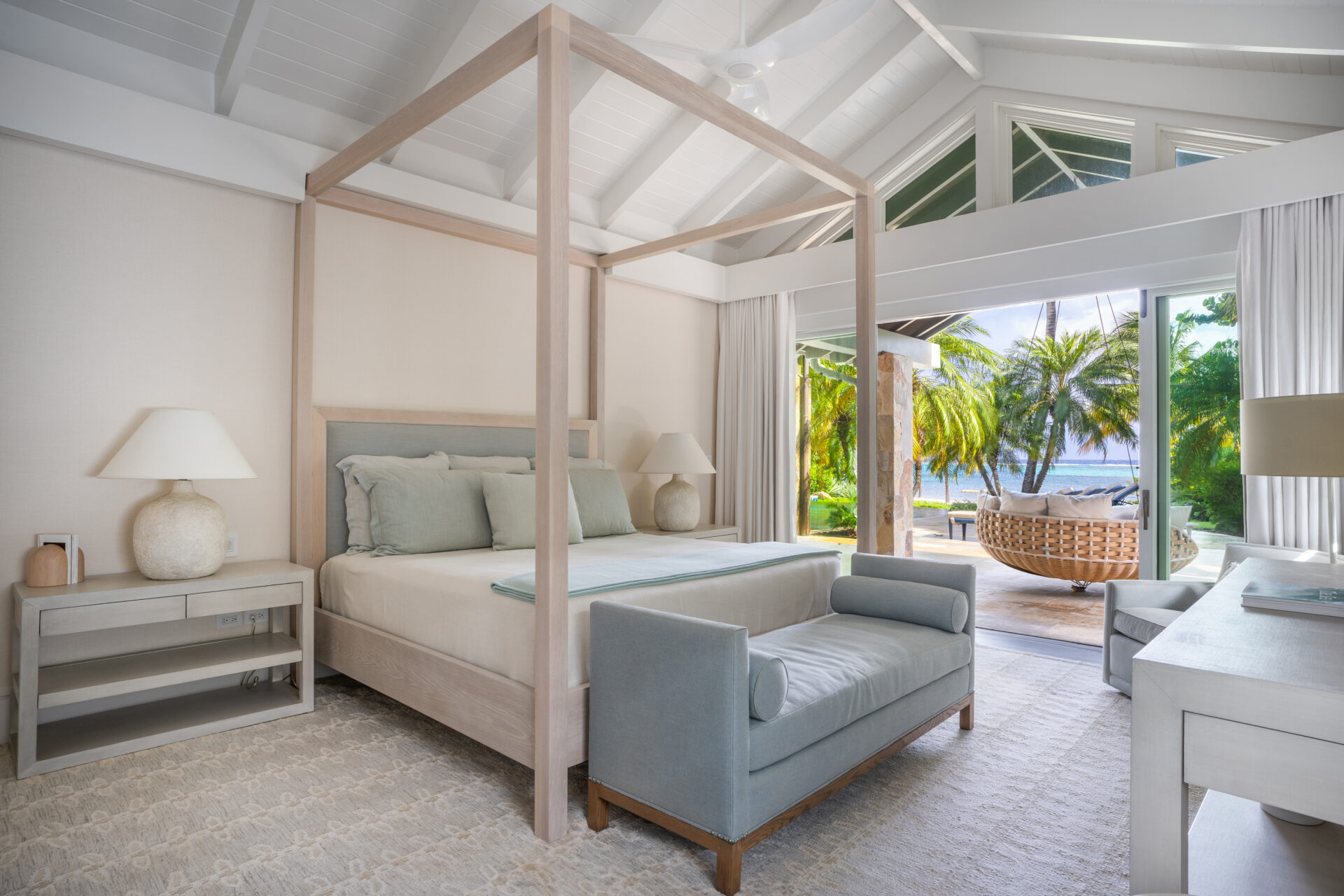 Welcome to Nonamé Villa, where a bright, airy bedroom features a canopy bed with light-colored linens and a cushioned bench at the foot. Large windows and sliding glass doors offer views of a tropical patio with wicker furniture, palm trees, and ocean scenery. The decor is clean and minimalistic.