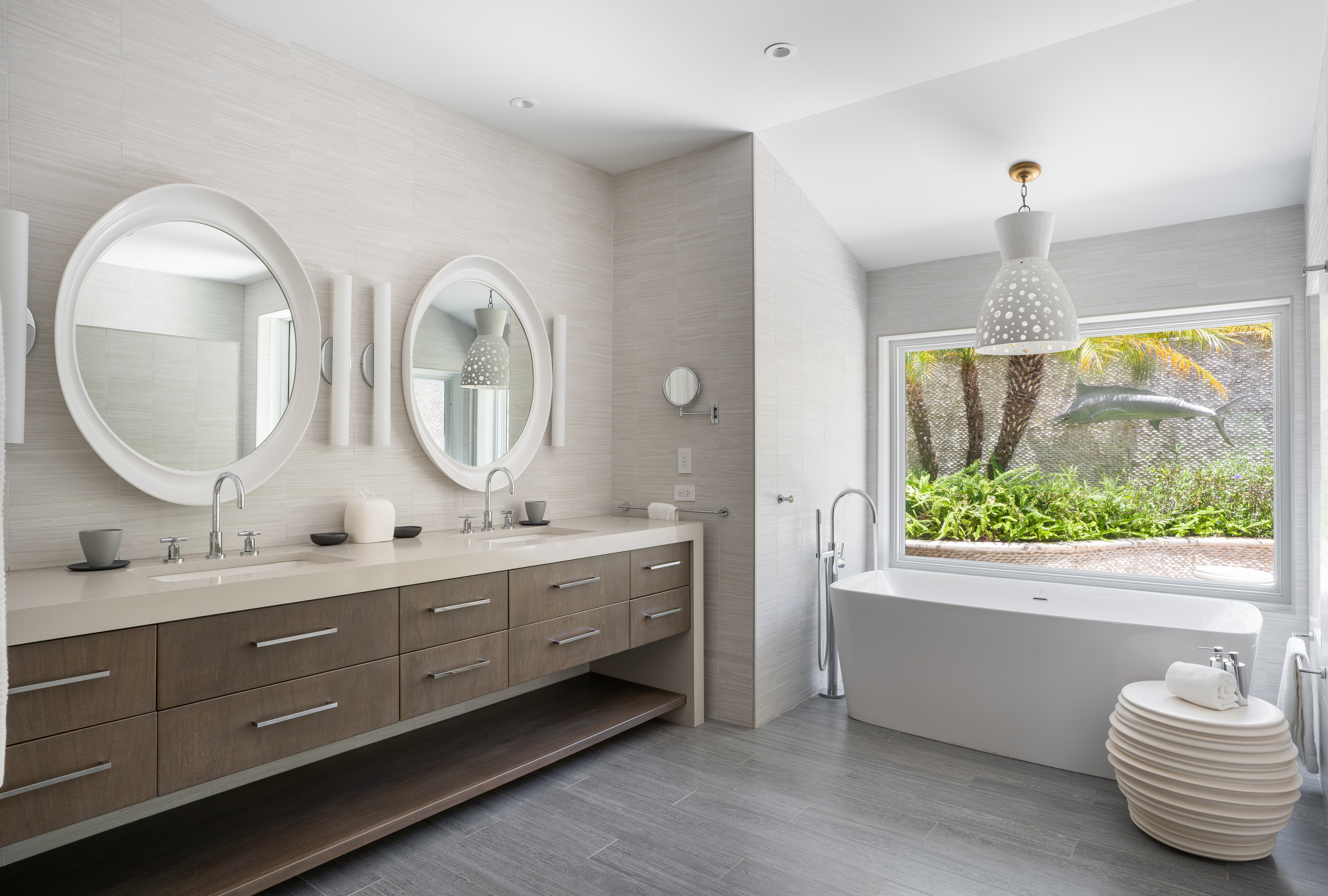 Modern bathroom at Nonamé Villa, featuring a double vanity with round mirrors, wall-mounted lights, and ample storage drawers. A freestanding bathtub is placed near a large window with views of a lush garden. Neutral tones and sleek design elements create a serene ambiance.