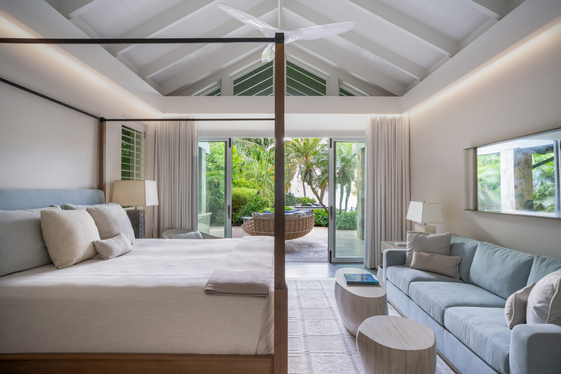 Spacious bedroom with high vaulted ceiling and exposed beams at Nonamé Villa. The room features a large canopy bed, a light blue sofa, and a coffee table. Glass doors open to a patio with outdoor seating, revealing lush greenery and palm trees outside. Neutral color scheme.