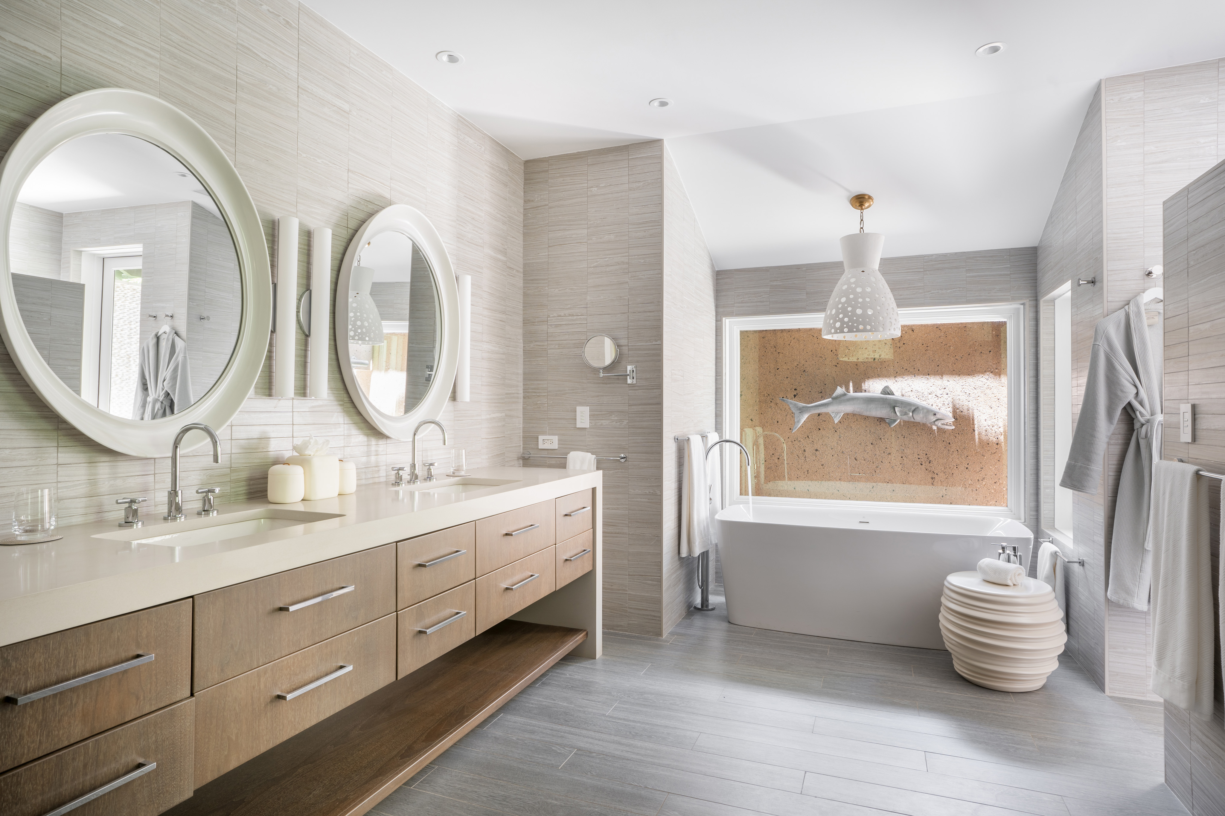 Modern bathroom in Nonamé Villa featuring a double vanity with two round mirrors, white countertops, and wooden cabinets. A freestanding bathtub is positioned in front of a decorative accent wall with a mounted fish art piece. The room has a clean, neutral color palette and ample lighting.