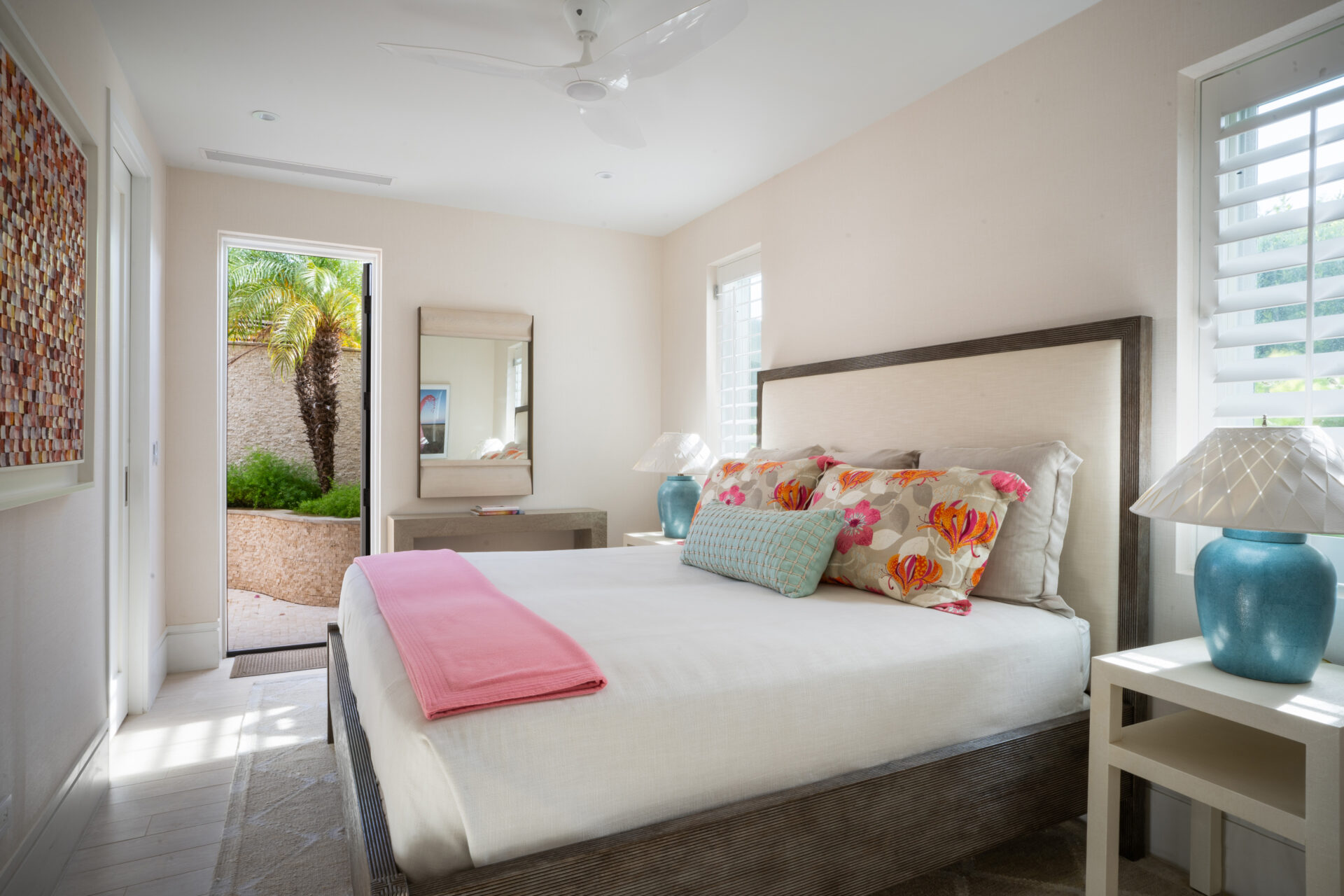 A bright bedroom at Nonamé Villa features a double bed with floral and pastel pillows, a pink blanket at the foot, and white bedding. The room has two bedside tables with blue lamps, a large framed art piece on the left wall, and a door leading to an outdoor patio with lush greenery.