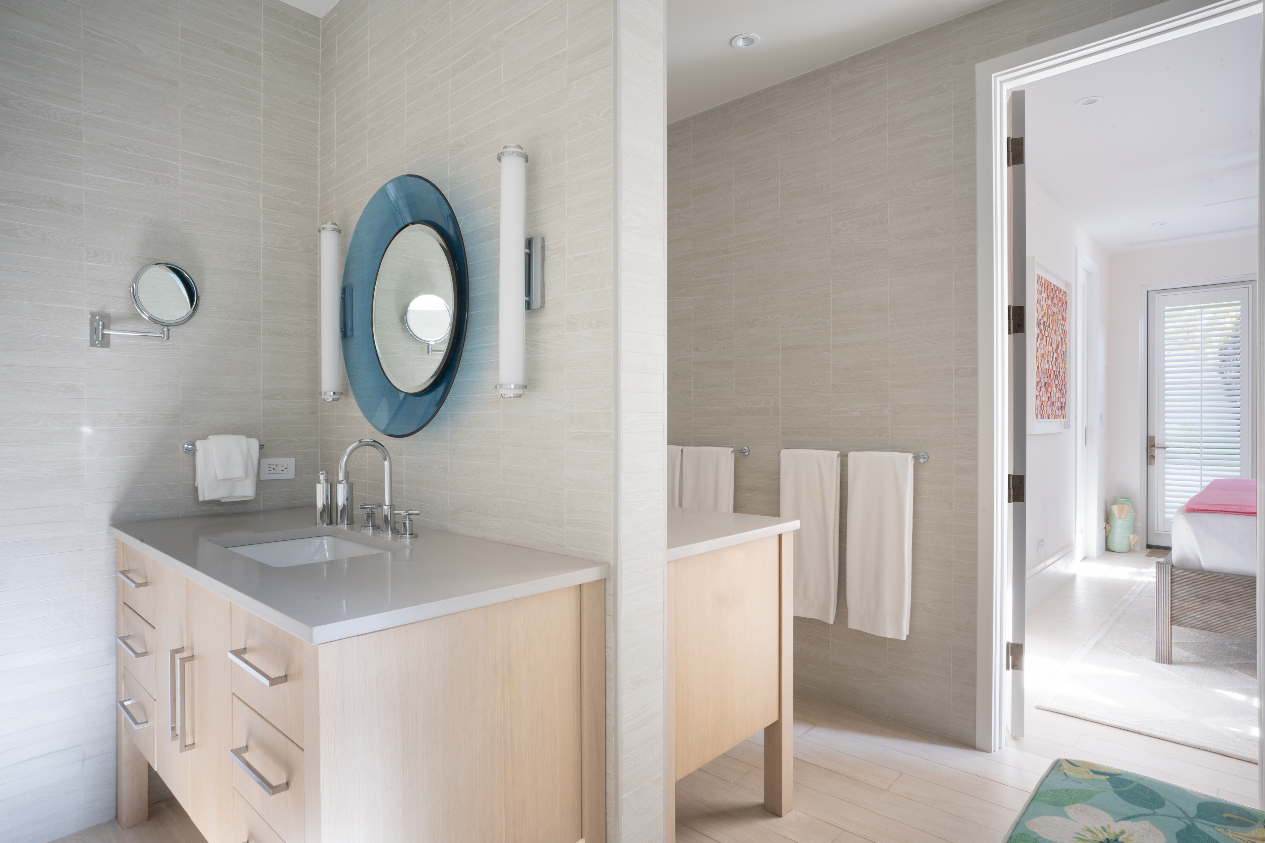 A modern bathroom at Nonamé Villa features a wooden vanity with a white countertop and an oval mirror. Wall-mounted lights flank the mirror. White towels hang on a rack, and through an open door, a glimpse of a bedroom with white walls and a colorful bedspread is visible.