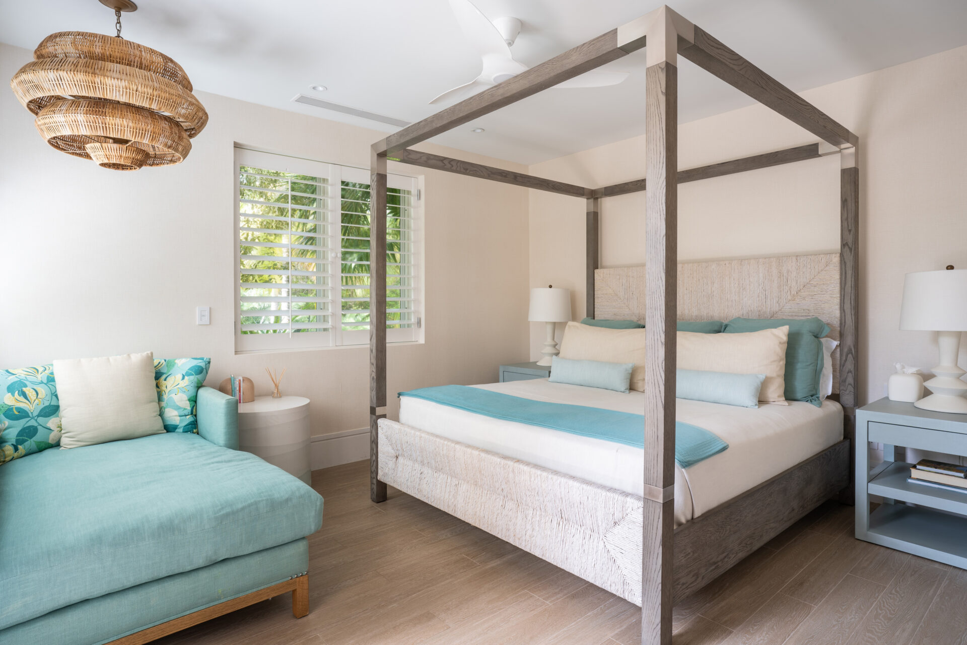 A bright bedroom in the Nonamé villa features a four-poster bed with white and light blue bedding, a teal chaise lounge with patterned cushions, and a modern ceiling fan. Natural light streams in through the window with white shutters, illuminating the light wood floor and neutral walls.