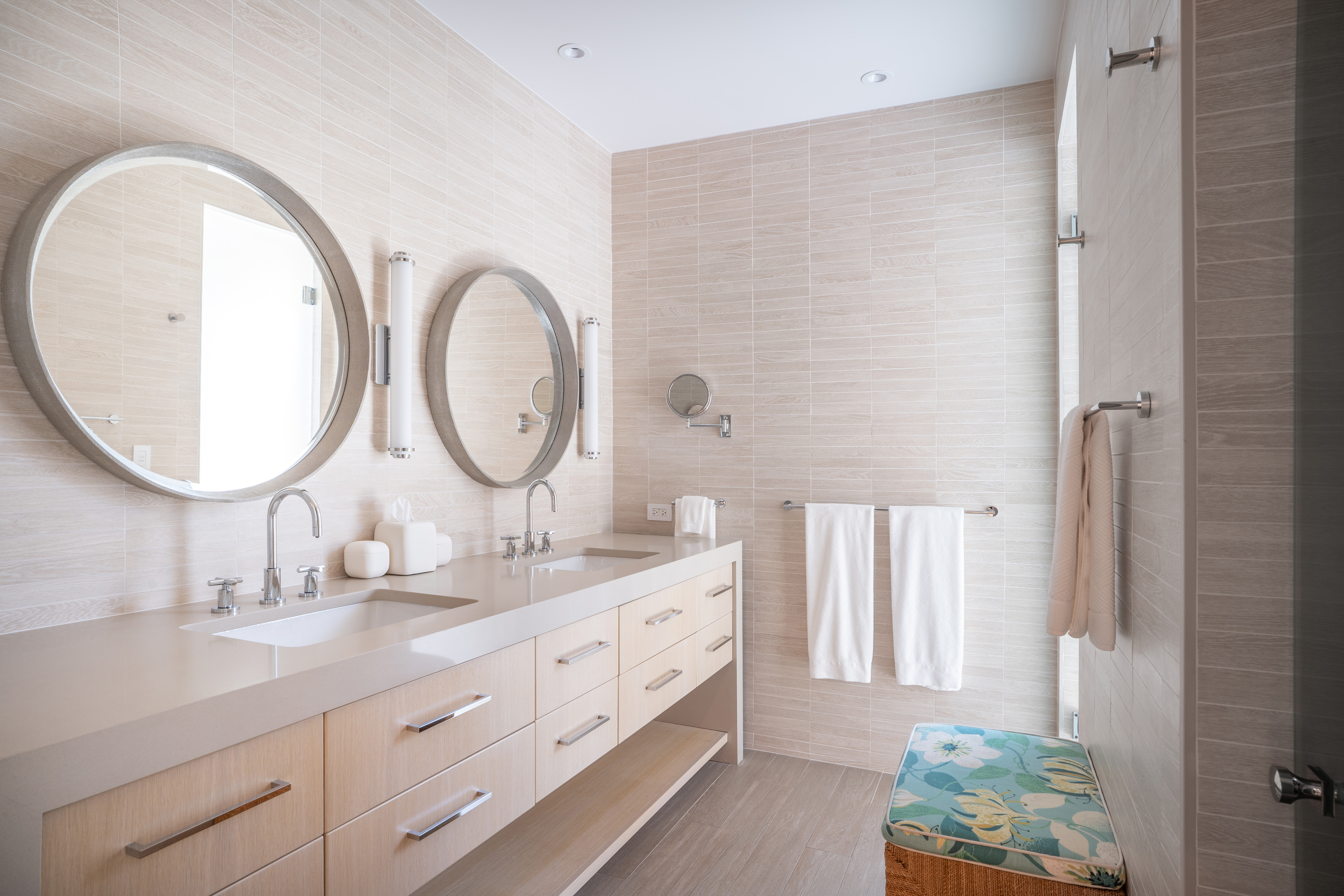 A modern bathroom at Nonamé Villa features light beige tiles and double sinks with large round mirrors and sleek faucets. White towels hang on a rack, and a wicker basket with a floral cushion is placed near a glass-enclosed shower. The space is well-lit with recessed lighting.