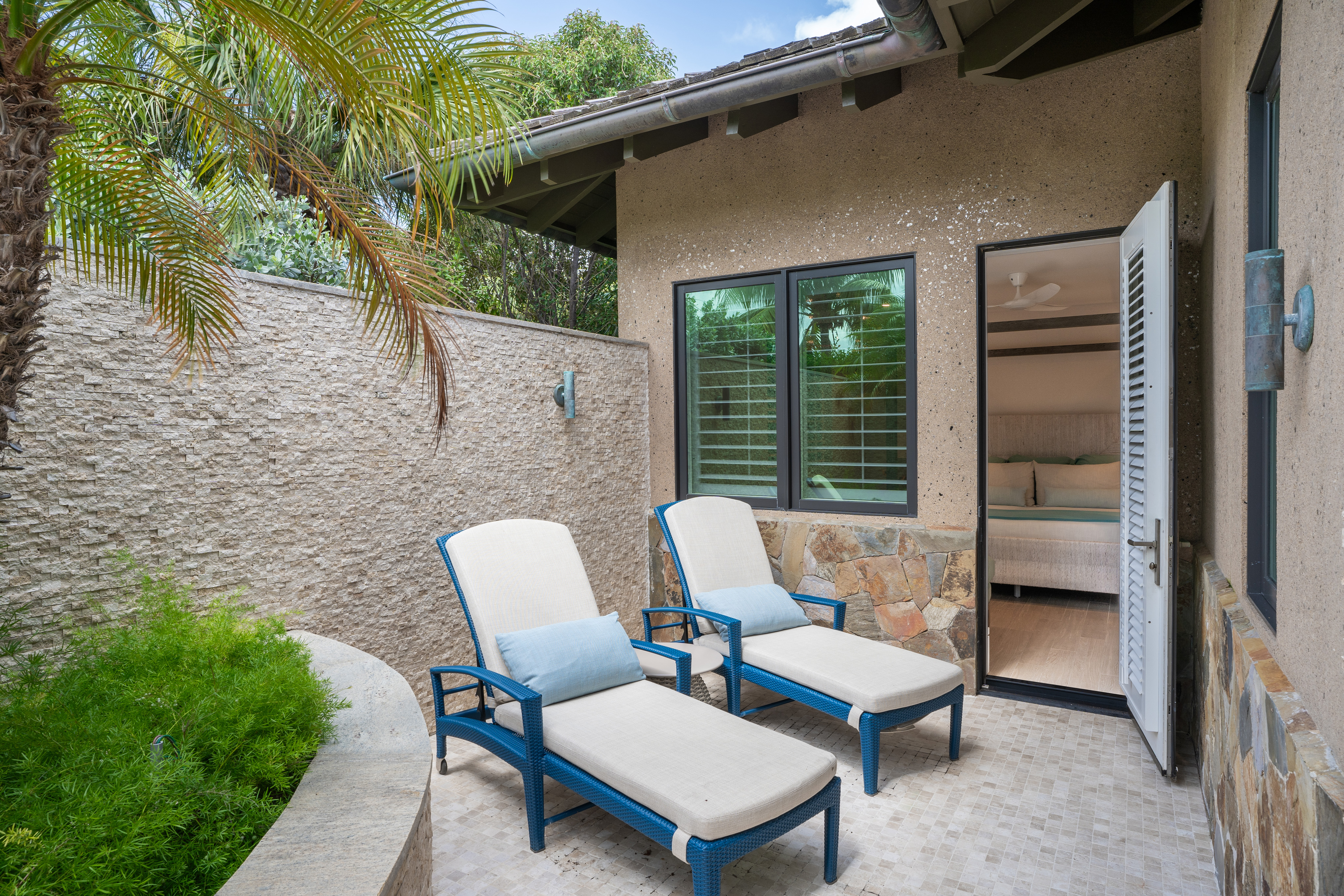 A cozy outdoor patio at Nonamé Villa features two blue metal lounge chairs with white cushions placed on a tiled floor. The patio is surrounded by a textured beige wall with greenery and a palm tree visible. An open door leads indoors, offering a glimpse of a bedroom.