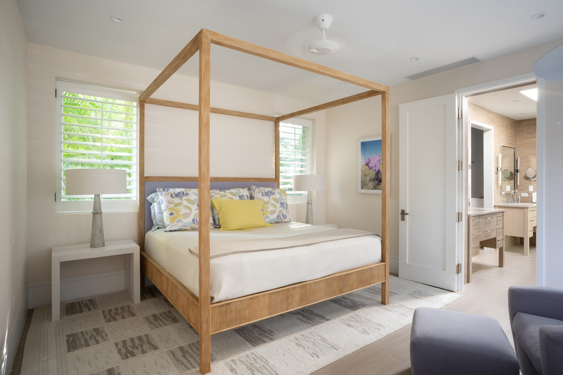 A modern, bright bedroom at Nonamé Villa features a wooden four-poster bed adorned with floral and yellow accent pillows. The room includes side tables with lamps, a large rug, and windows with white shutters. An open door leads to an airy bathroom with a double vanity and modern fixtures.