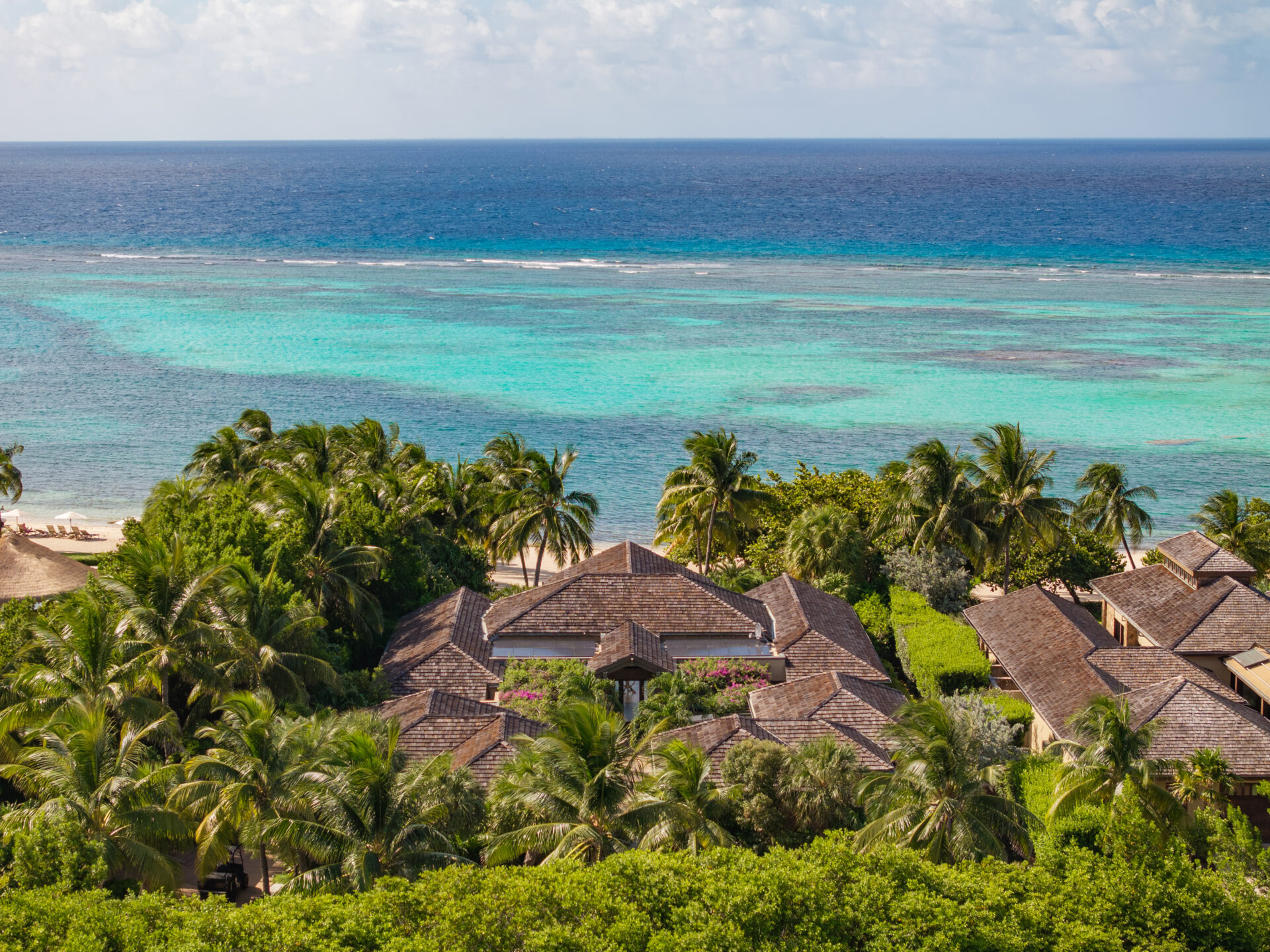 A scenic view of Nonamé Villa, a tropical resort featuring multiple thatched-roof buildings nestled among lush green palm trees. In the background, there is a vibrant turquoise ocean under a partly cloudy sky. The shoreline is visible with sand stretching along the coast.