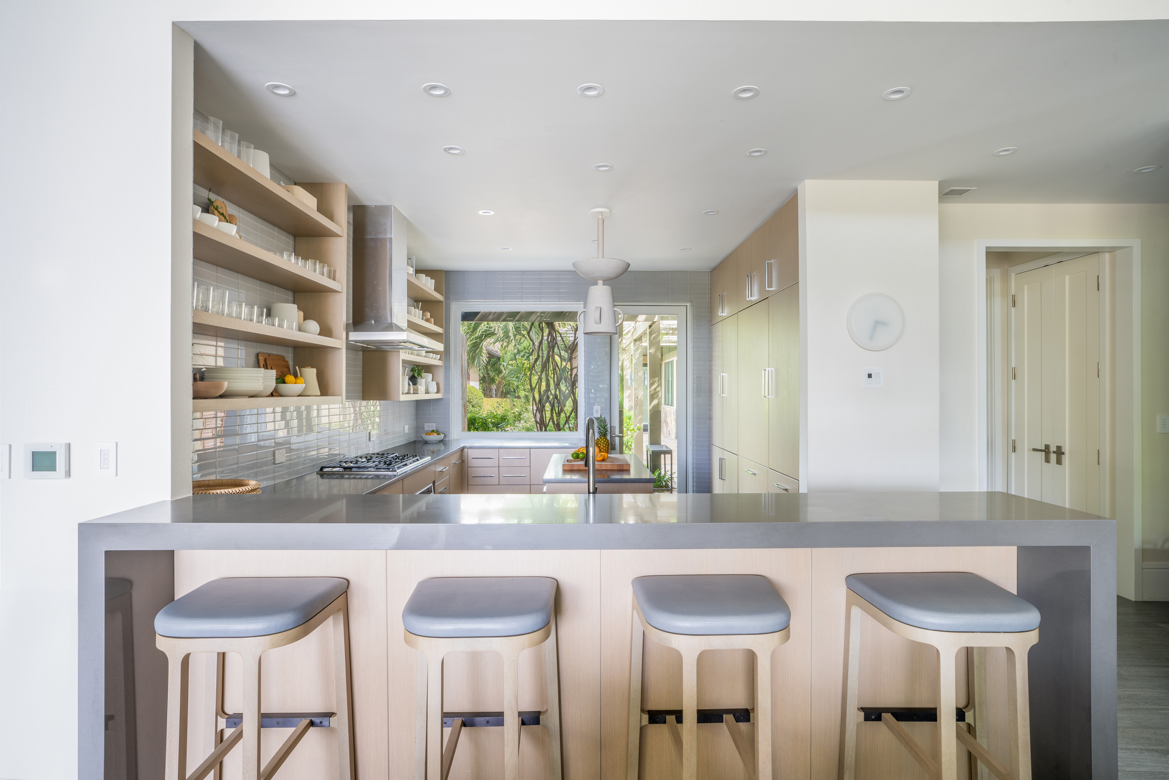 The modern kitchen at Nonamé Villa boasts a minimalist design, featuring a long, grey countertop with four light wood stools. Open shelving displays dishes and glassware, while a large window offers a view of greenery outside. The space is bright with natural light and clean lines.