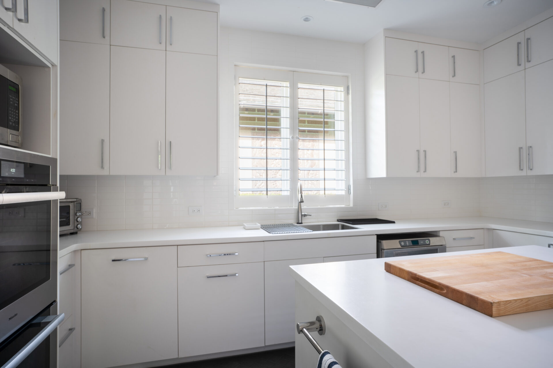 A modern kitchen at Nonamé Villa features white cabinetry, drawers, and countertops. A wooden cutting board graces the island, while stainless steel appliances, including an oven, microwave, and dishwasher, are visible. A window above the sink lets in natural light.