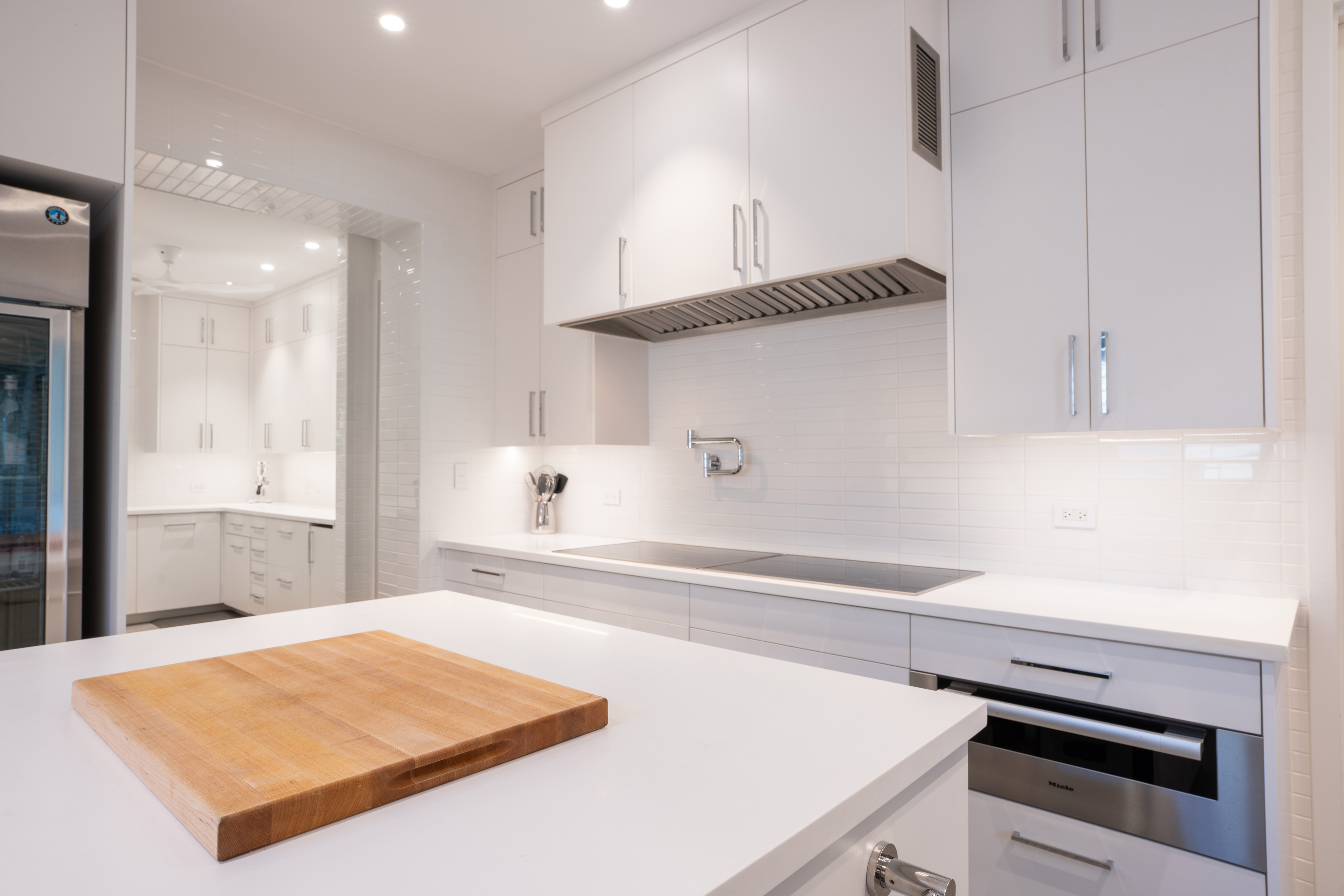 A modern kitchen in Nonamé Villa features white cabinets, countertops, and backsplash. An induction cooktop is built into the counter with a stainless steel hood above. A wooden cutting board rests on the center island. The space is well-lit with recessed ceiling lights.