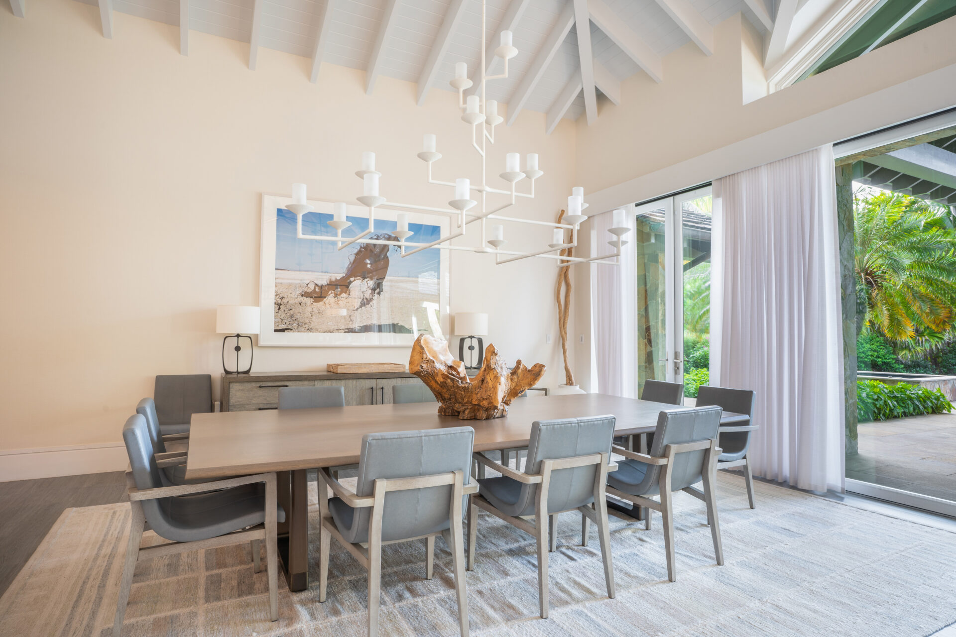 Noname Villa boasts a spacious dining room with a vaulted ceiling, featuring a rectangular wooden table surrounded by eight gray cushioned chairs. A modern chandelier hangs above, and large windows offer views of greenery outside. A large framed beach scene decorates the back wall.