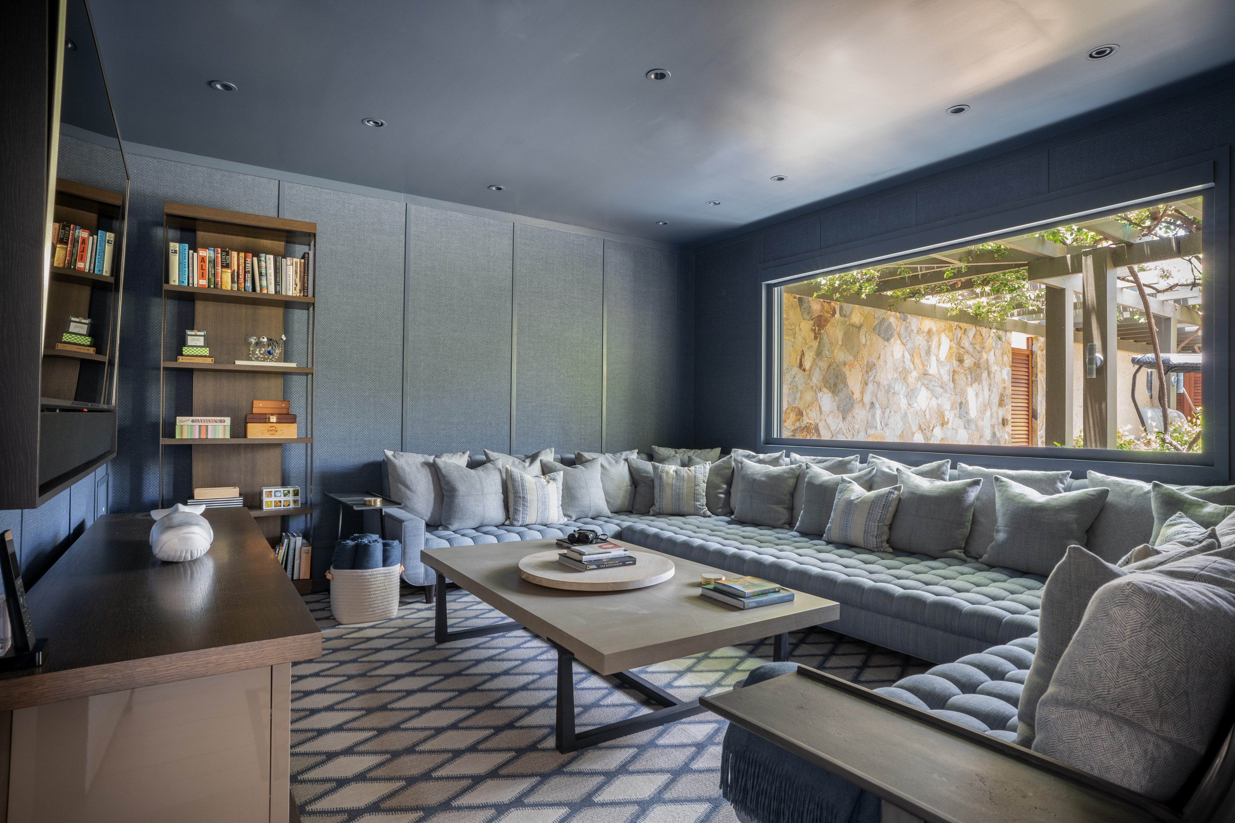 A cozy living room at Nonamé Villa features a large sectional sofa filled with numerous cushions. A modern coffee table sits in the center, flanked by open bookshelves filled with books and decorative items. A large window offers a view of a stone wall and lush greenery outside.