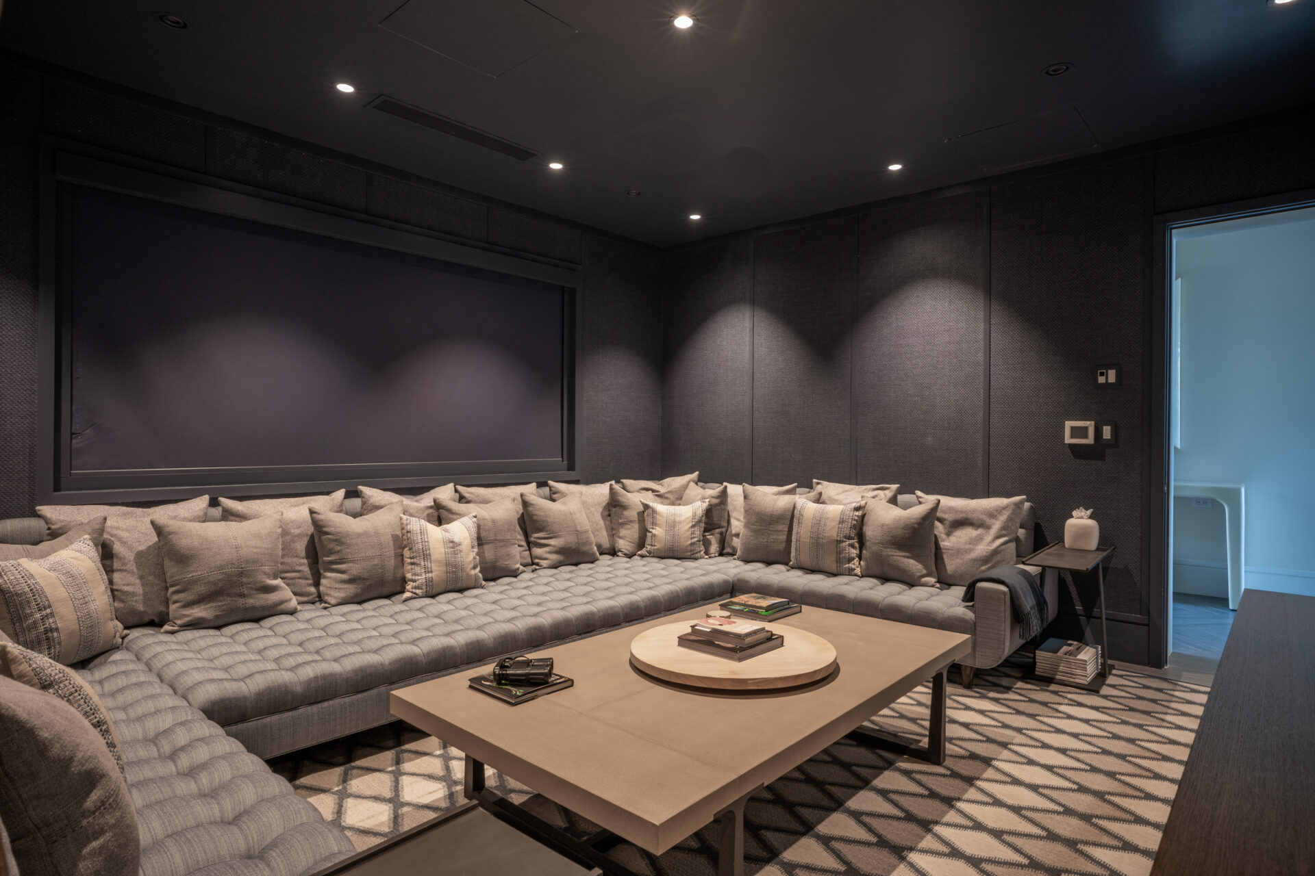 A modern, dimly lit living room in Nonamé Villa features a large U-shaped gray sofa adorned with multiple gray pillows. In front of the sofa is a wooden coffee table with a few books on it. The room has dark walls and lighting from recessed ceiling lights.