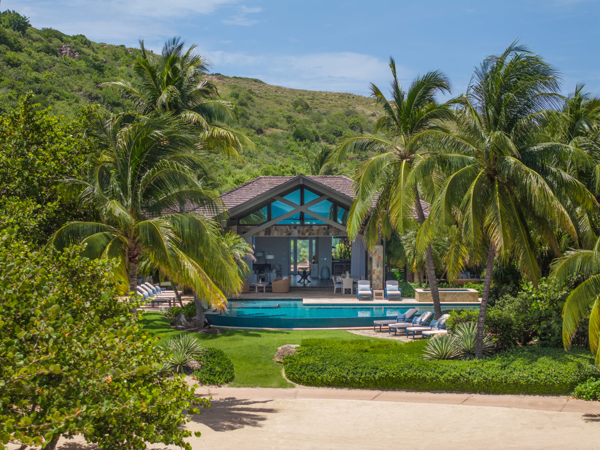 Nonamé Villa is a luxurious beachfront retreat with a sleek modern design, surrounded by lush palm trees and greenery. The villa features a large outdoor pool with sun loungers and an open-air living space under a gabled roof, set against the backdrop of a gently sloping, forested hill.