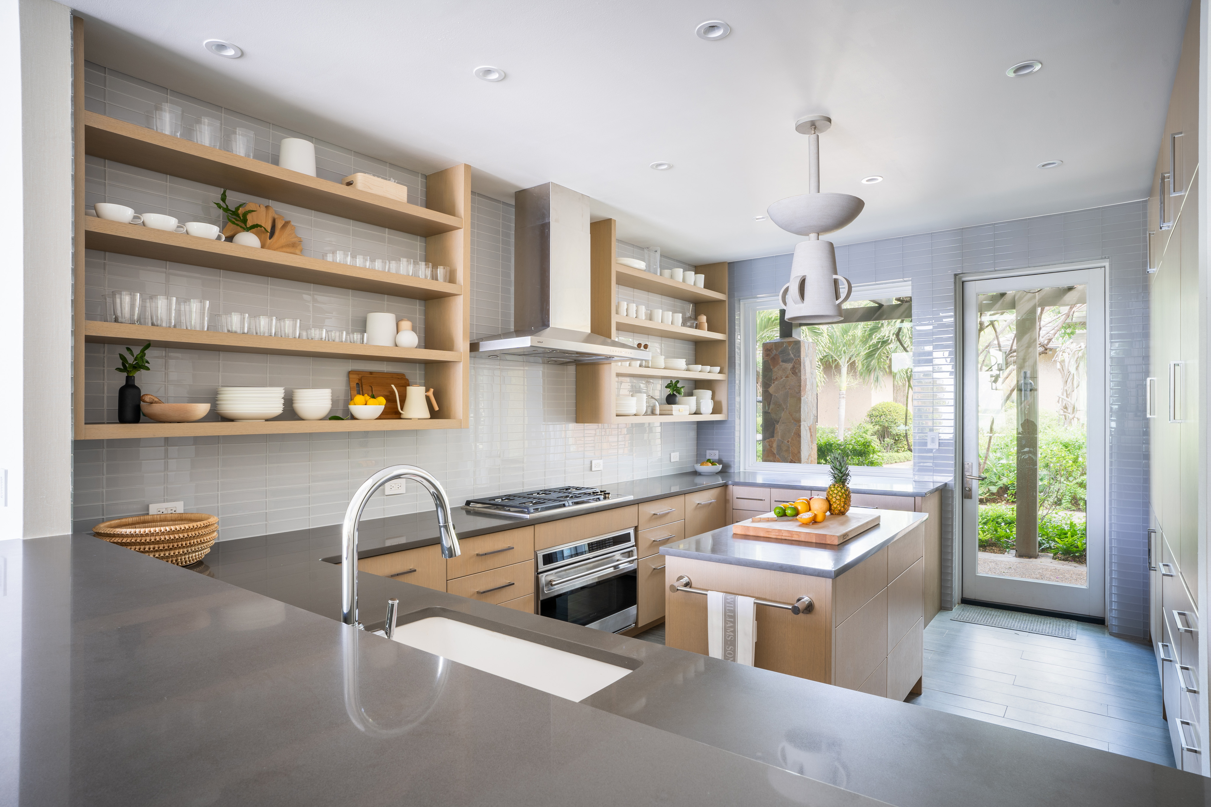 Modern kitchen in a villa with light wood cabinets, open shelving displaying dishes, and stainless steel appliances. A large island with a fruit bowl and cutting board sits in the center, under a contemporary Nonamé pendant light. A door and window offer a view of a lush garden.