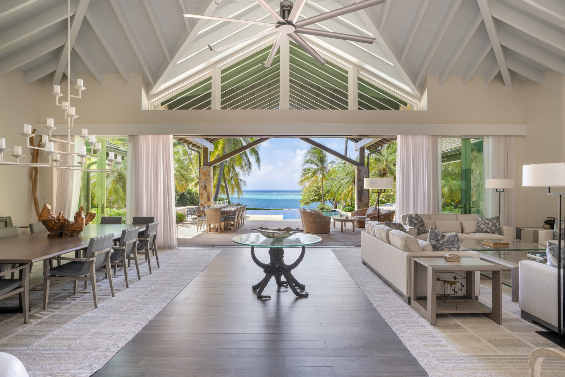 Spacious, modern open-concept living and dining area with high cathedral ceilings, large windows, and glass doors leading to a patio. The view extends to a tropical beach with palm trees and the ocean. At Nonamé Villa, a large round table sits in the center of the room.