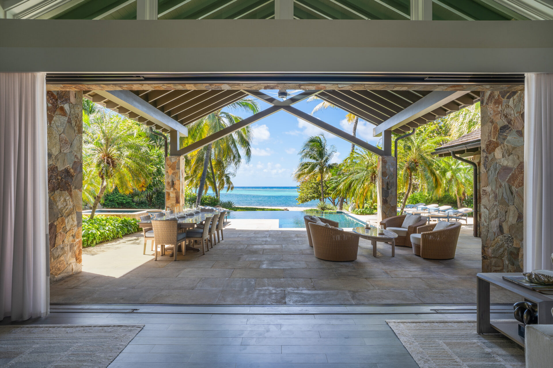 The Nonamé Villa boasts an open-air living space that leads to a patio with outdoor seating and dining areas, featuring wicker furniture. Beyond the patio, a pool overlooks a scenic ocean view framed by palm trees under a clear blue sky. The space is partially covered with a vaulted roof.