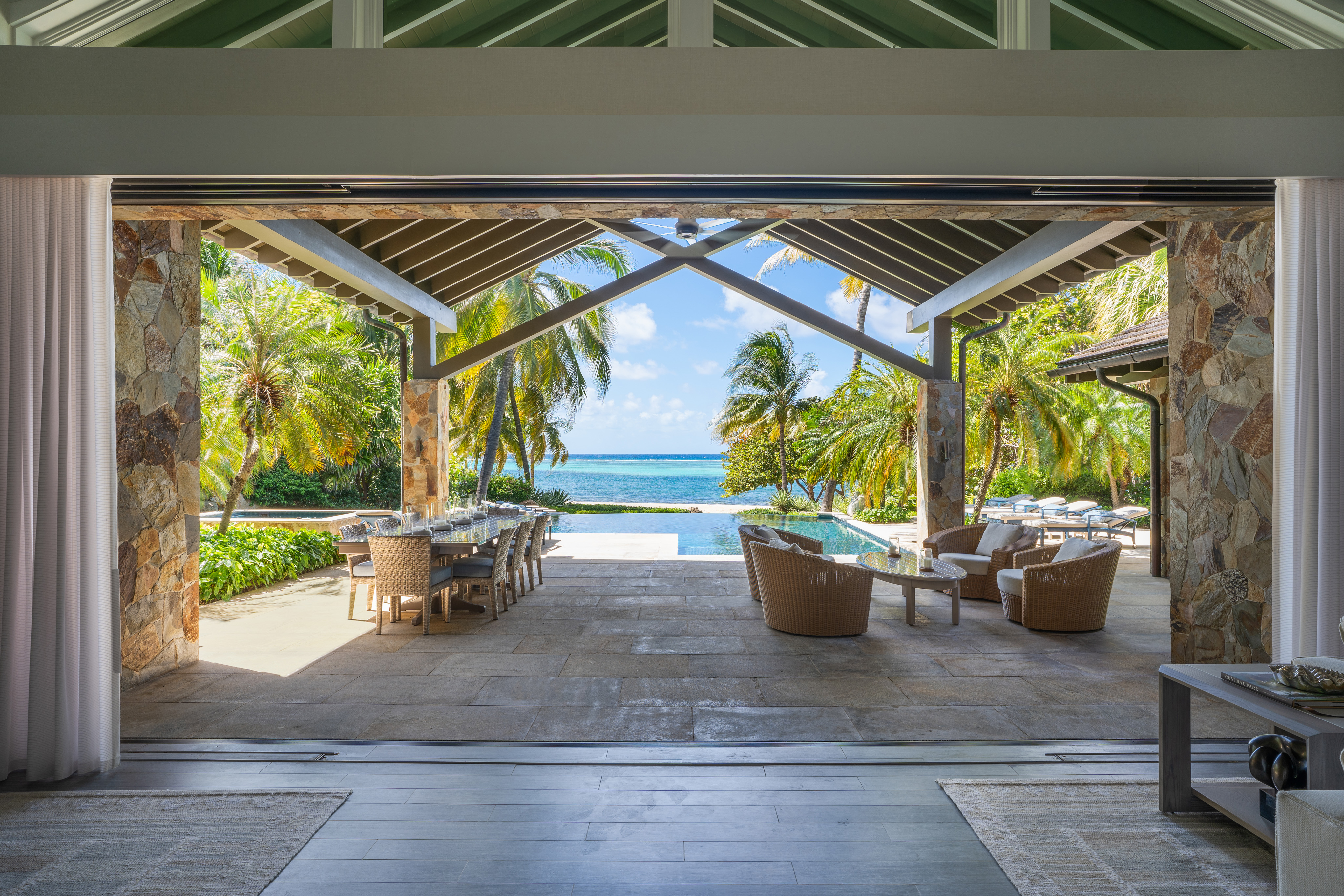 The Nonamé Villa boasts an open-air living space that leads to a patio with outdoor seating and dining areas, featuring wicker furniture. Beyond the patio, a pool overlooks a scenic ocean view framed by palm trees under a clear blue sky. The space is partially covered with a vaulted roof.