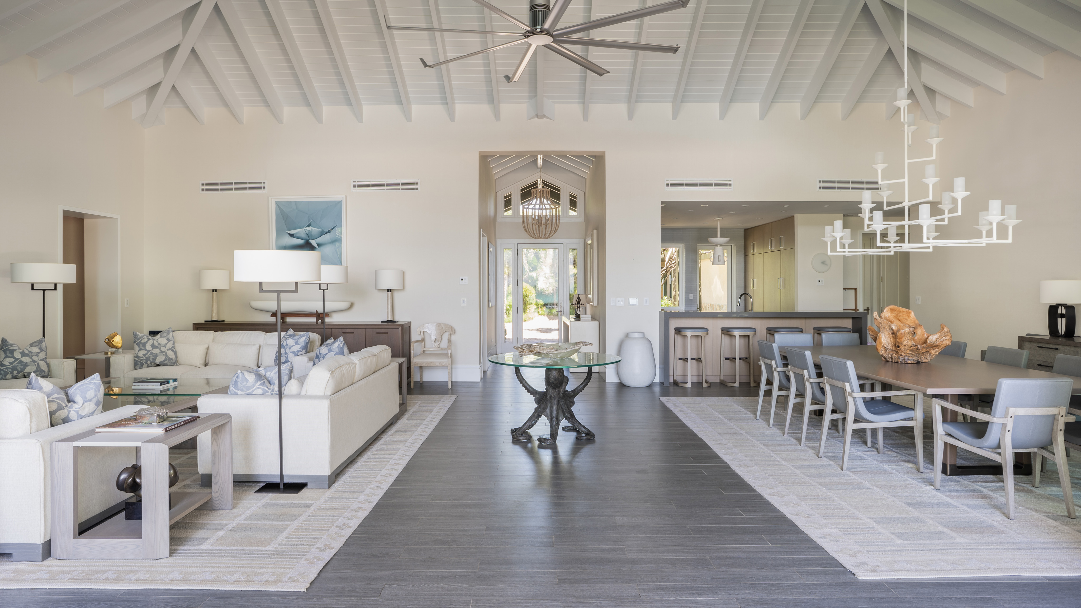 Spacious, airy living and dining area at Nonamé Villa with light-colored furniture, high vaulted ceiling, and modern decor. Features a large ceiling fan, chandeliers, and floor-to-ceiling windows overlooking a scenic outdoor view. Open kitchen with bar stools in the background.