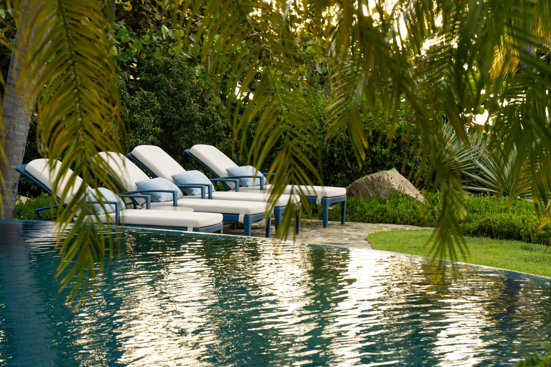 Four white cushioned lounge chairs are lined up alongside a pool at Nonamé Villa, surrounded by lush greenery and palm fronds. The serene setting suggests a relaxing atmosphere, perfect for lounging and enjoying the peaceful outdoor environment.