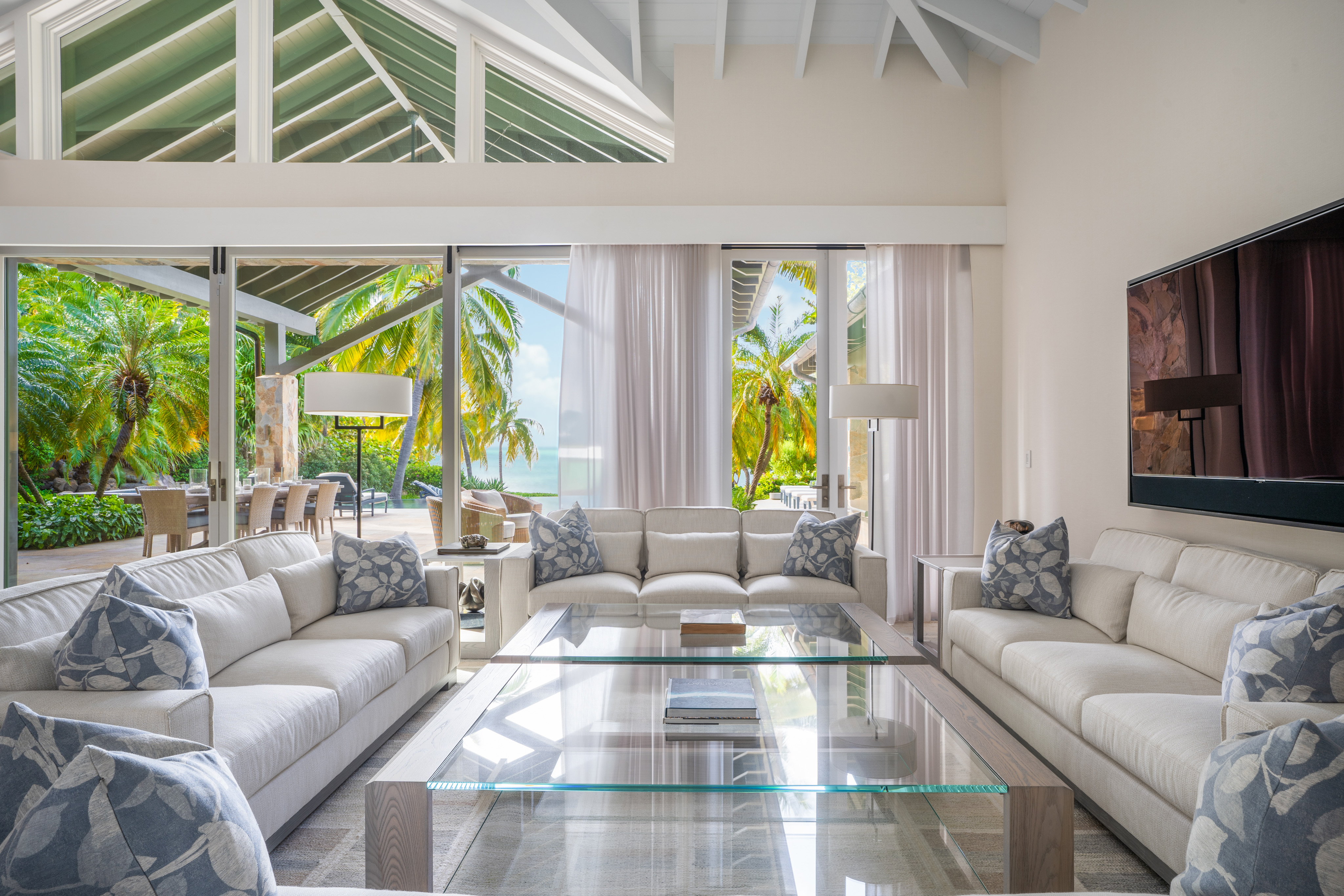 Spacious and bright living room at Nonamé Villa with large glass windows offering a view of a tropical outdoor area. The room features white sofas arranged around a glass coffee table with decorative cushions. A high ceiling with exposed beams adds to the airy feel.