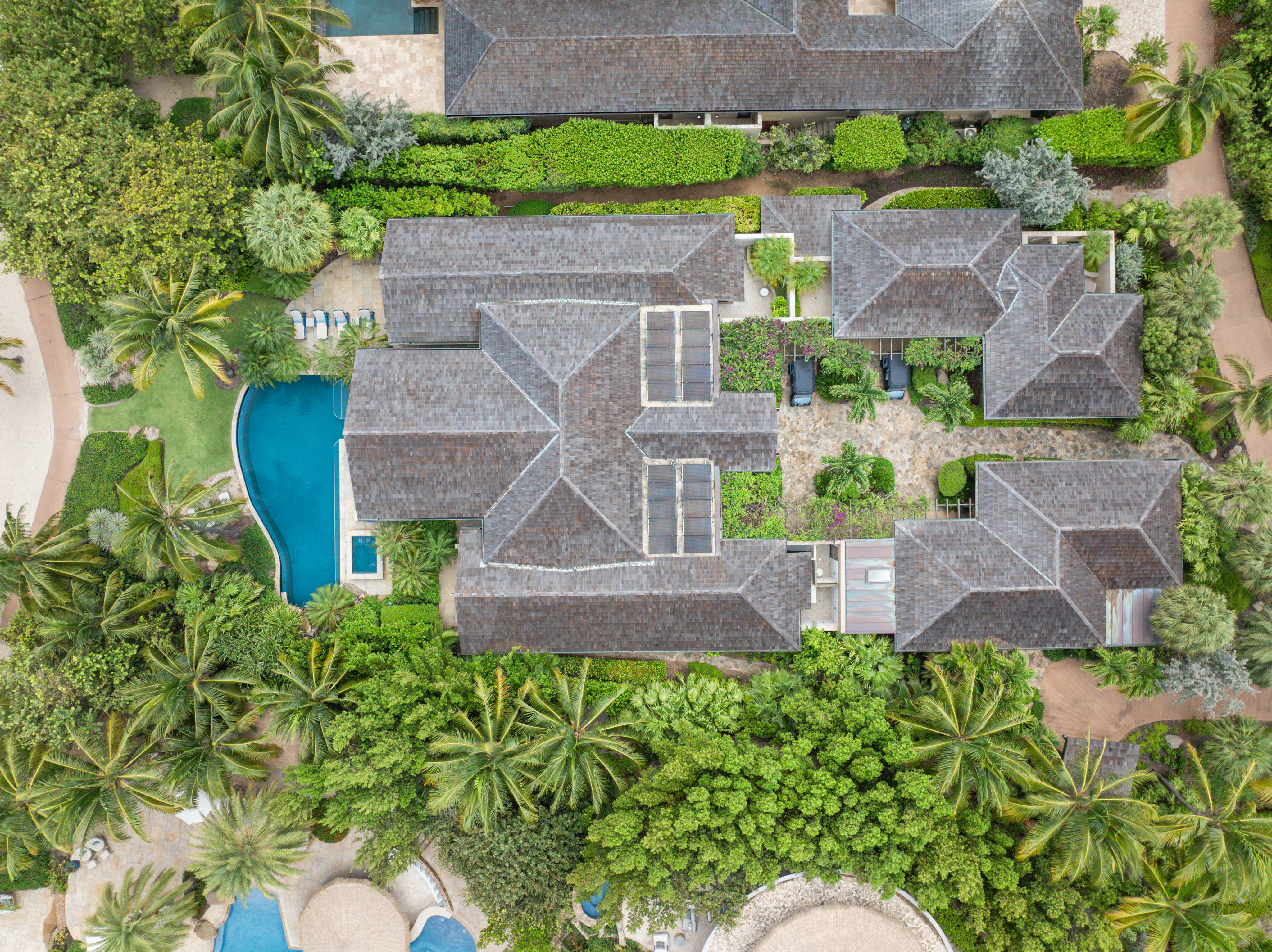 Aerial view of Nonamé Villa, a large, luxurious estate with multiple buildings, lush greenery, and a swimming pool. The house has a distinct, complex roof design. The property is surrounded by tall trees, blending into a tropical landscape. Paths and gardens are visible throughout.